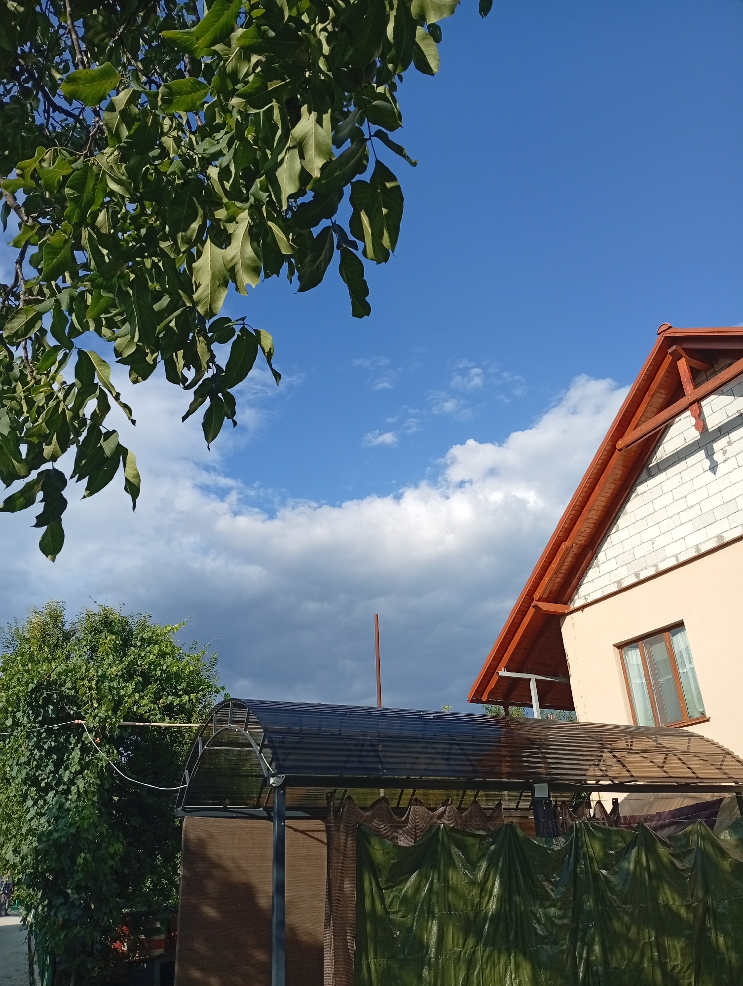 The image depicts an outdoor scene with a clear blue sky, partially obscured by fluffy white and darker gray clouds. In the foreground, a tree with lush green leaves extends into the upper left corner of the frame. To the right, a portion of a house is visible, featuring a light peach-colored wall and a dark red-tiled roof with exposed wooden beams. A single window with a brown frame and white curtains is set into the wall. Below the house and extending across the middle ground is a covered structure with a translucent dark roof supported by a metal frame. This structure is draped with a green tarp or fabric in the lower part of the image, and a brown screen is visible behind it. To the left of this covered area, a dense green tree is present, and a thin white wire runs horizontally across the scene. The overall impression is that of a residential property in a semi-rural or suburban setting.