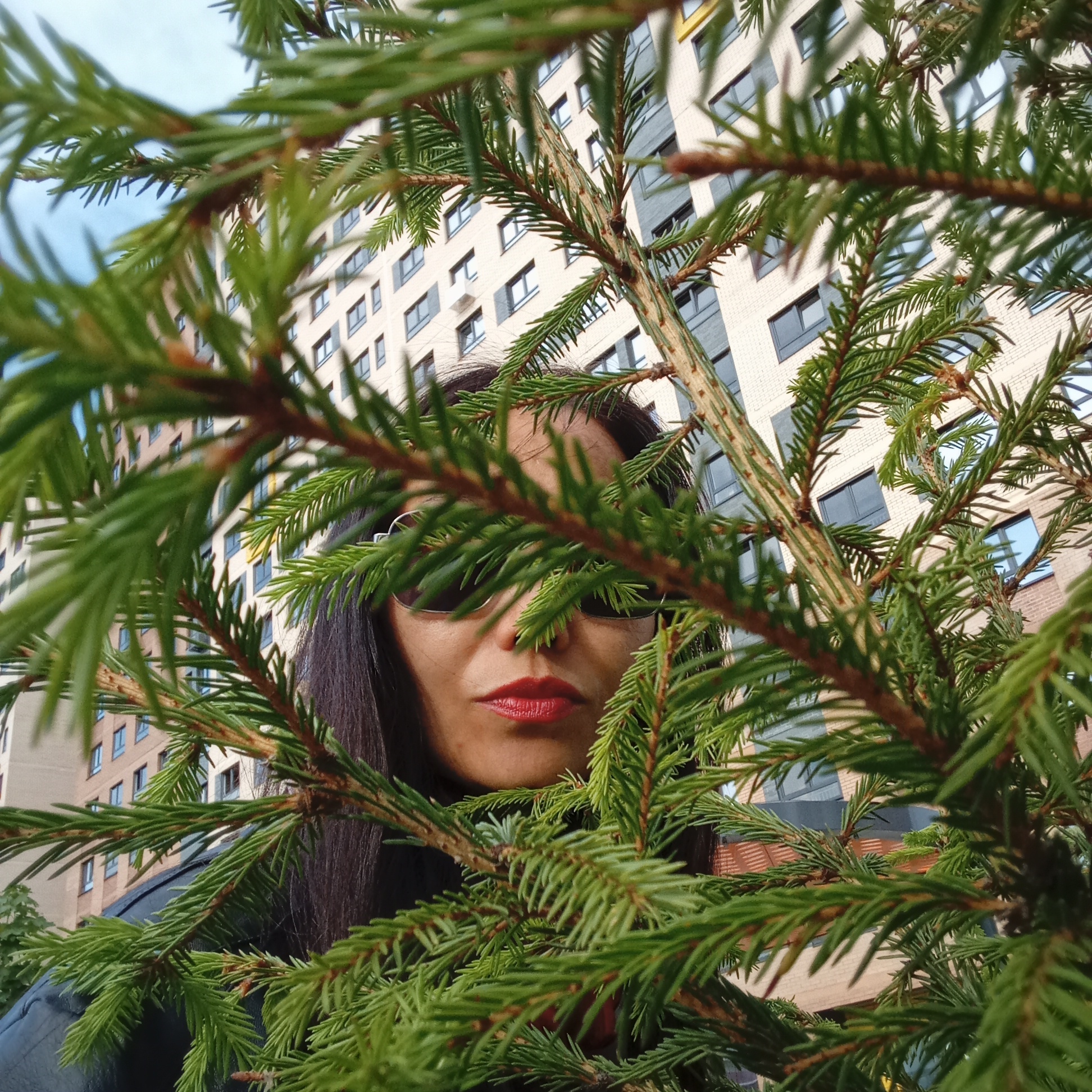 The image presents a close-up, eye-level portrait of a woman partially framed by vibrant green evergreen branches. The woman has long, dark hair and is wearing dark sunglasses, obscuring her eyes. Her lips are painted with prominent red lipstick. Only her face, parts of her hair, and shoulders are visible as she peeks through the dense foliage, looking directly towards the viewer.
In the background, a large, modern, multi-story residential building is visible under a clear or lightly overcast sky. The building features a light-colored facade, possibly brick or concrete, with numerous dark, rectangular windows arranged in rows. The architectural style suggests an urban or suburban setting, consistent with the location context of Izhevsk, Russia.
The scene is set during the daytime, indicated by the bright, even lighting and visible sky. The evergreen branches, likely from a spruce or fir tree, are sharply in focus in the foreground, providing a natural, organic frame for the woman's face. The activity captured is a posed portrait, with the branches adding an element of depth and mystery. There is no visible text in the image.