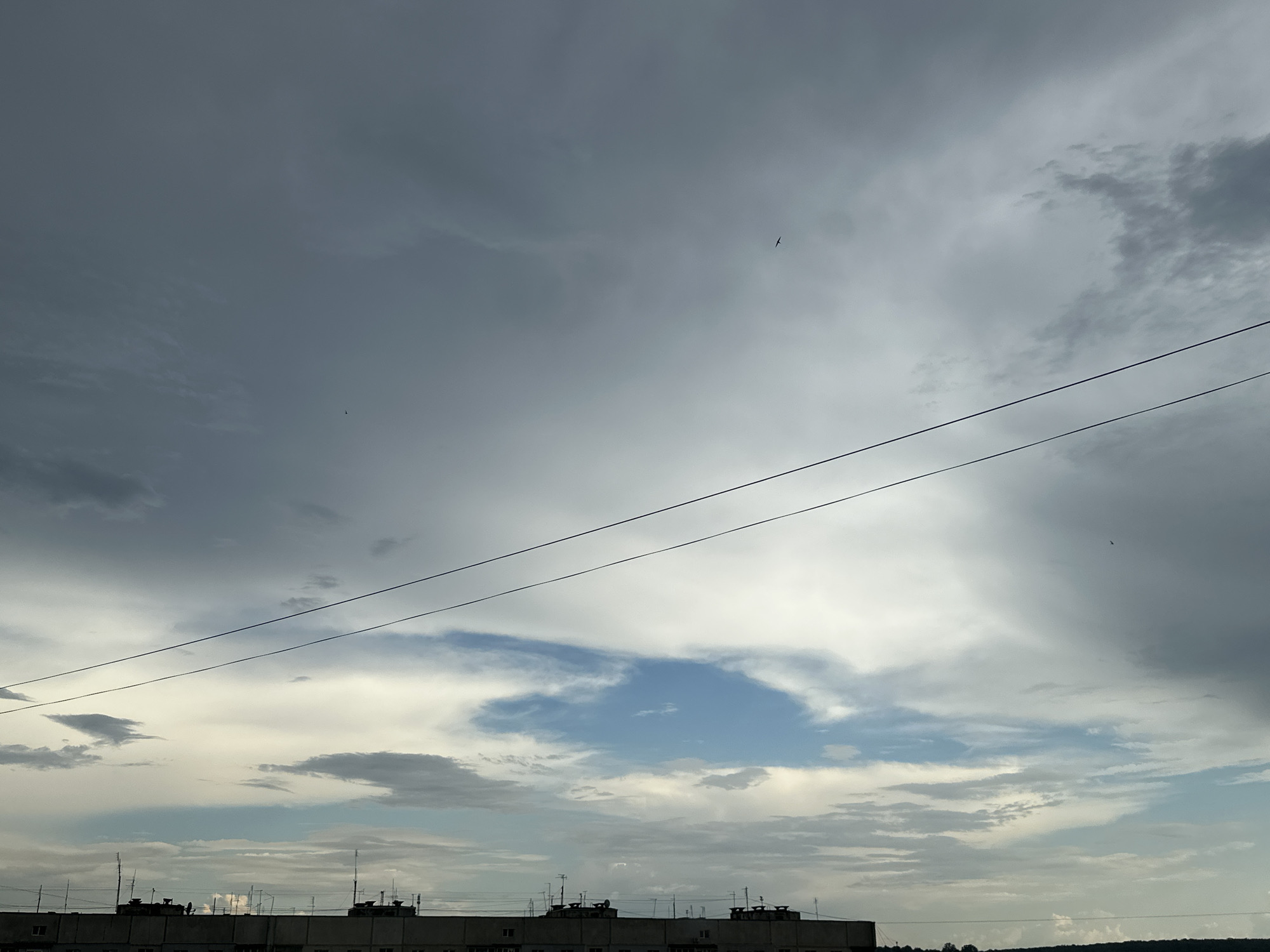A wide shot shows a cloudy sky above a city skyline. The sky is filled with various types of clouds, from dark, heavy storm clouds on the left to lighter, fluffy clouds on the right, with patches of blue sky visible in the center. Two dark power lines stretch horizontally across the frame, bisecting the sky. In the lower portion of the image, the tops of buildings are visible, forming a silhouette against the sky. These buildings appear to be apartment blocks or similar structures, and they are adorned with numerous antennas and other rooftop equipment. The overall impression is of an urban environment under a dynamic sky.