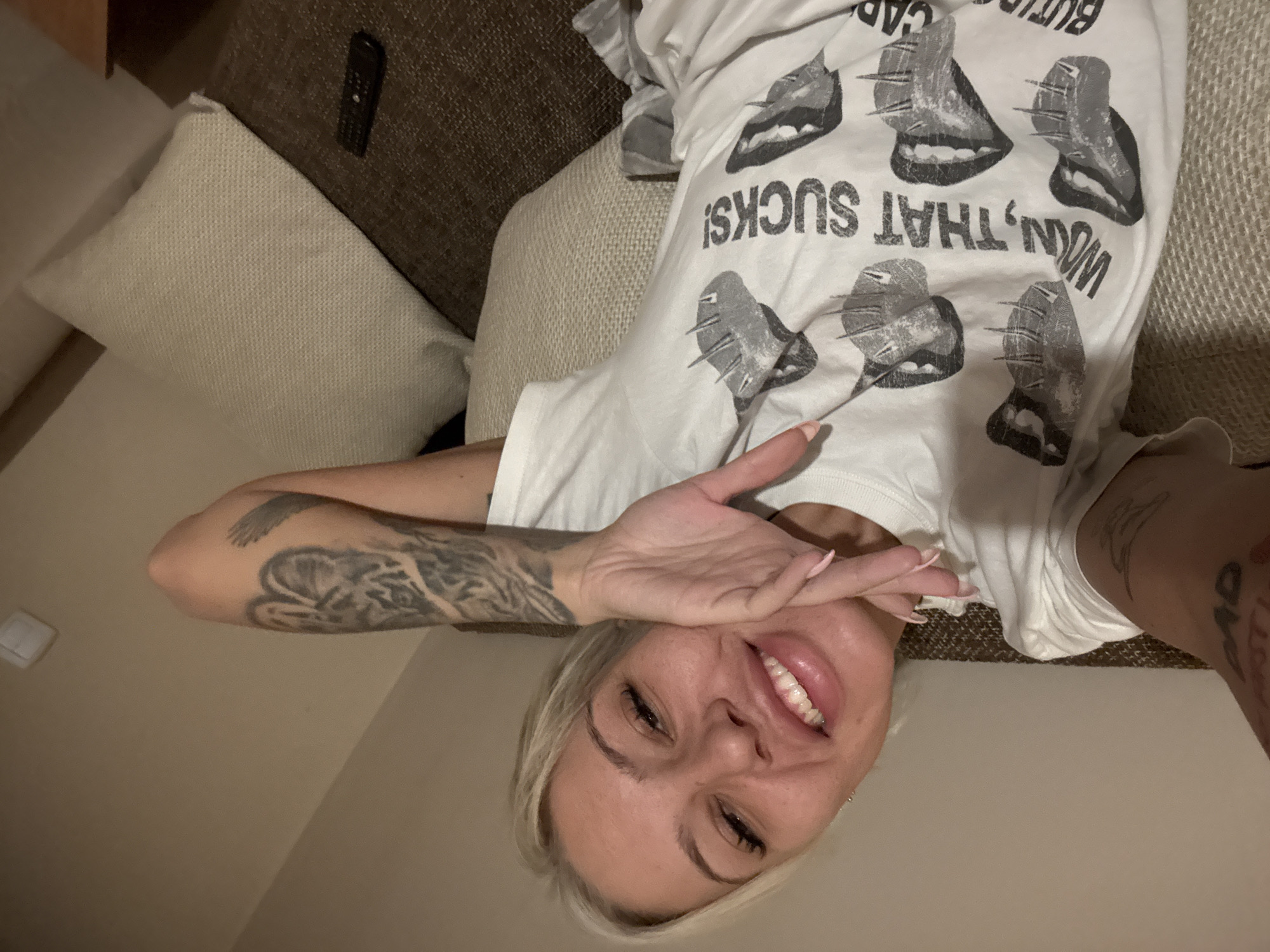 The image shows a woman lying on a couch. She has blonde hair and is wearing a white t-shirt with a graphic design of lips and the phrase "WOM, THAT SUCKS!". She is smiling and covering her eye with her hand. The couch is upholstered in a light brown fabric with some pillows. A remote control is also visible on the couch. The location is unknown and there's no evidence to suggest it is in Plovdiv, Bulgaria.