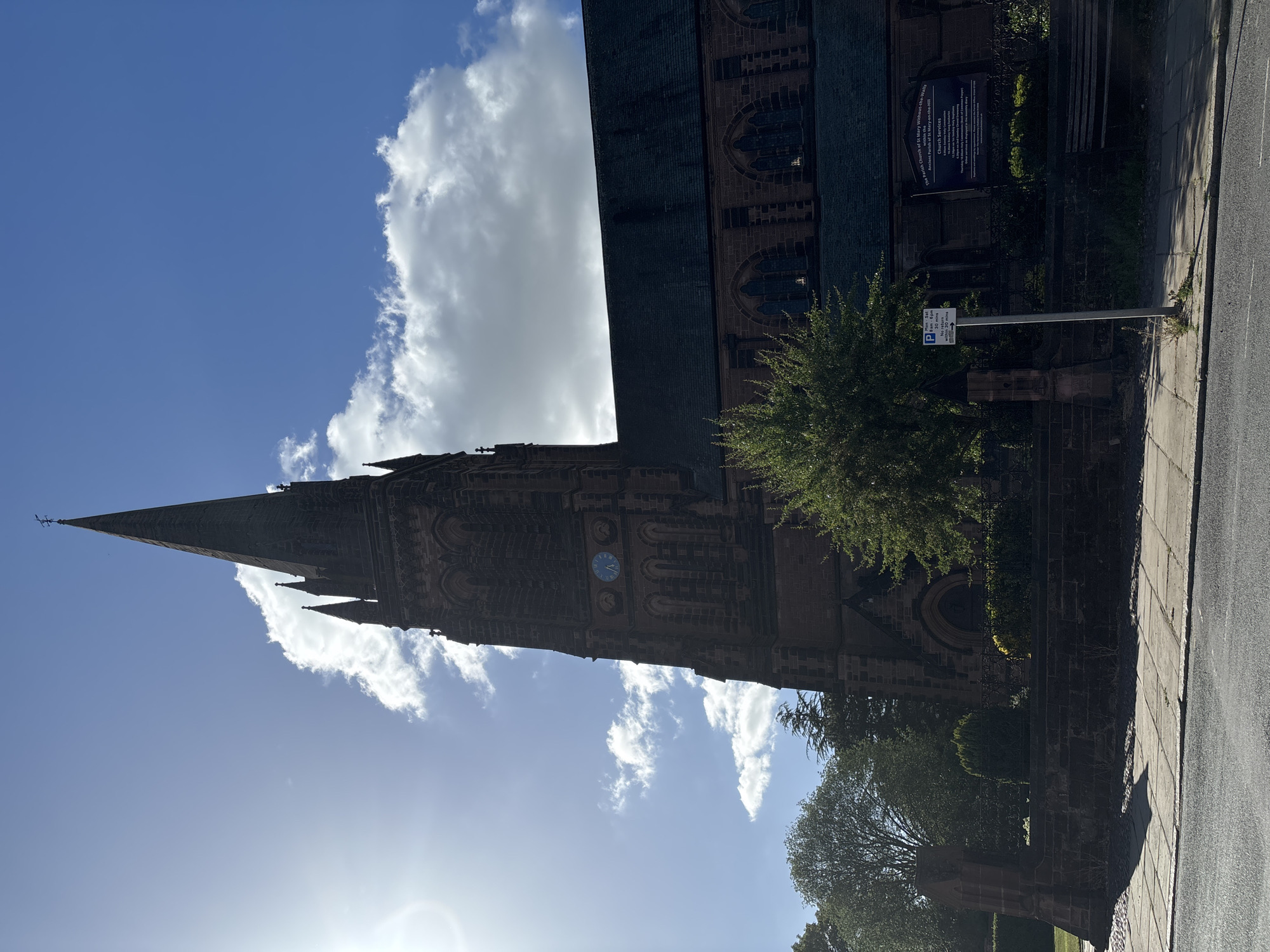 The image captures a view of The Parish Church of St Mary Without-the-Walls, located in Chester, United Kingdom. The church's steeple rises prominently against a blue sky dotted with white clouds. Architectural details of the stone structure, including arched windows and decorative elements, are visible. A clock is visible on the tower. Alongside the church, a section of the sidewalk and a nearby street can be seen. A parking sign and a notice board titled "The Parish Church of St Mary Without-the-Walls Ancient Parish of St Mary-on-the-Hill Church Services" stand adjacent to the building. Trees and greenery are present around the church.