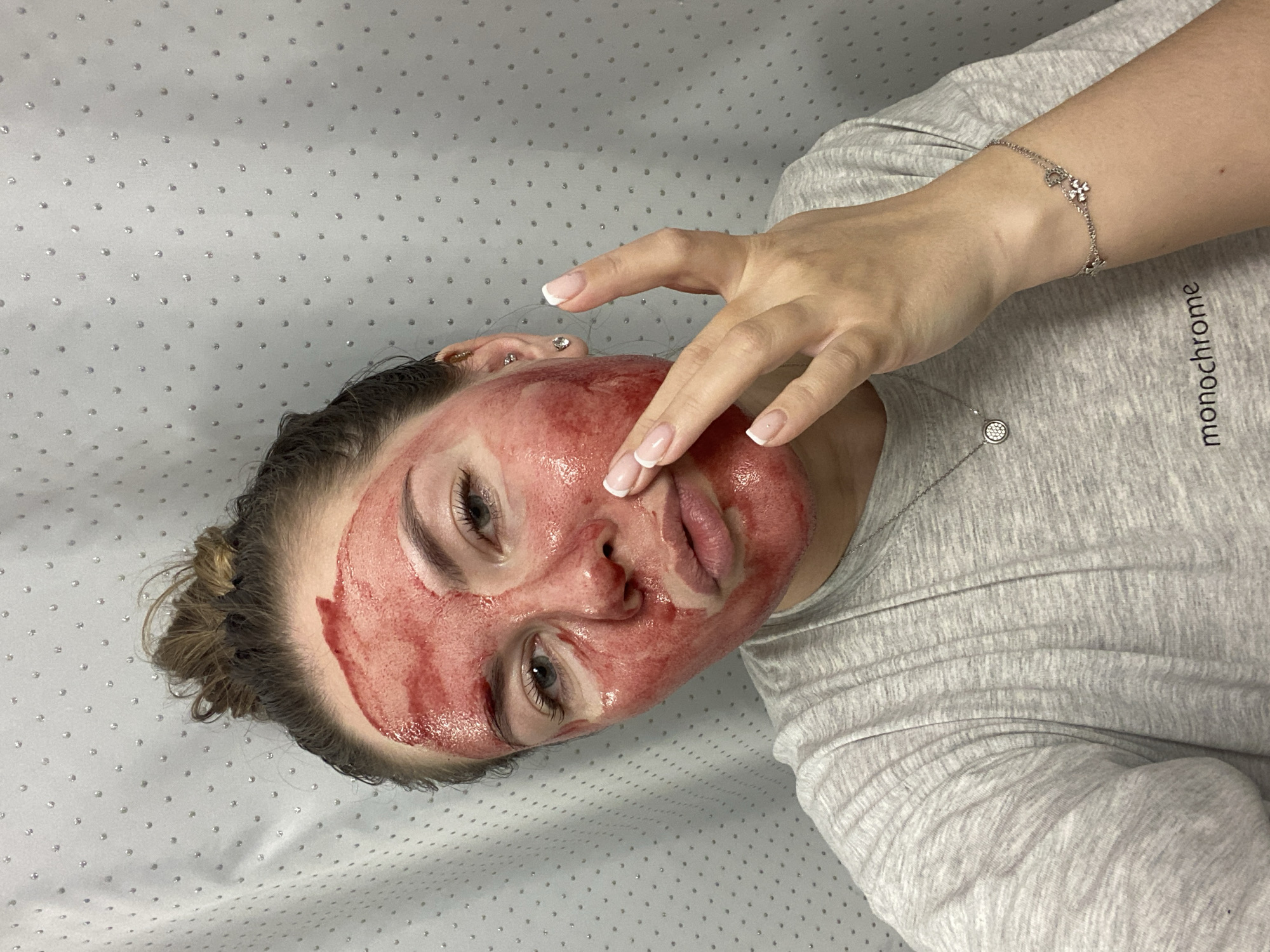A woman is lying on her back on a white surface with small, evenly spaced silver dots. She is wearing a light grey t-shirt. There is a silver bracelet on her wrist. Her face is covered in a red substance, possibly a facial treatment or blood. She is touching her face with her right hand. The name "monochrome" is printed on her shirt. The location is unknown and there is no indication of Izhevsk, Russia.
