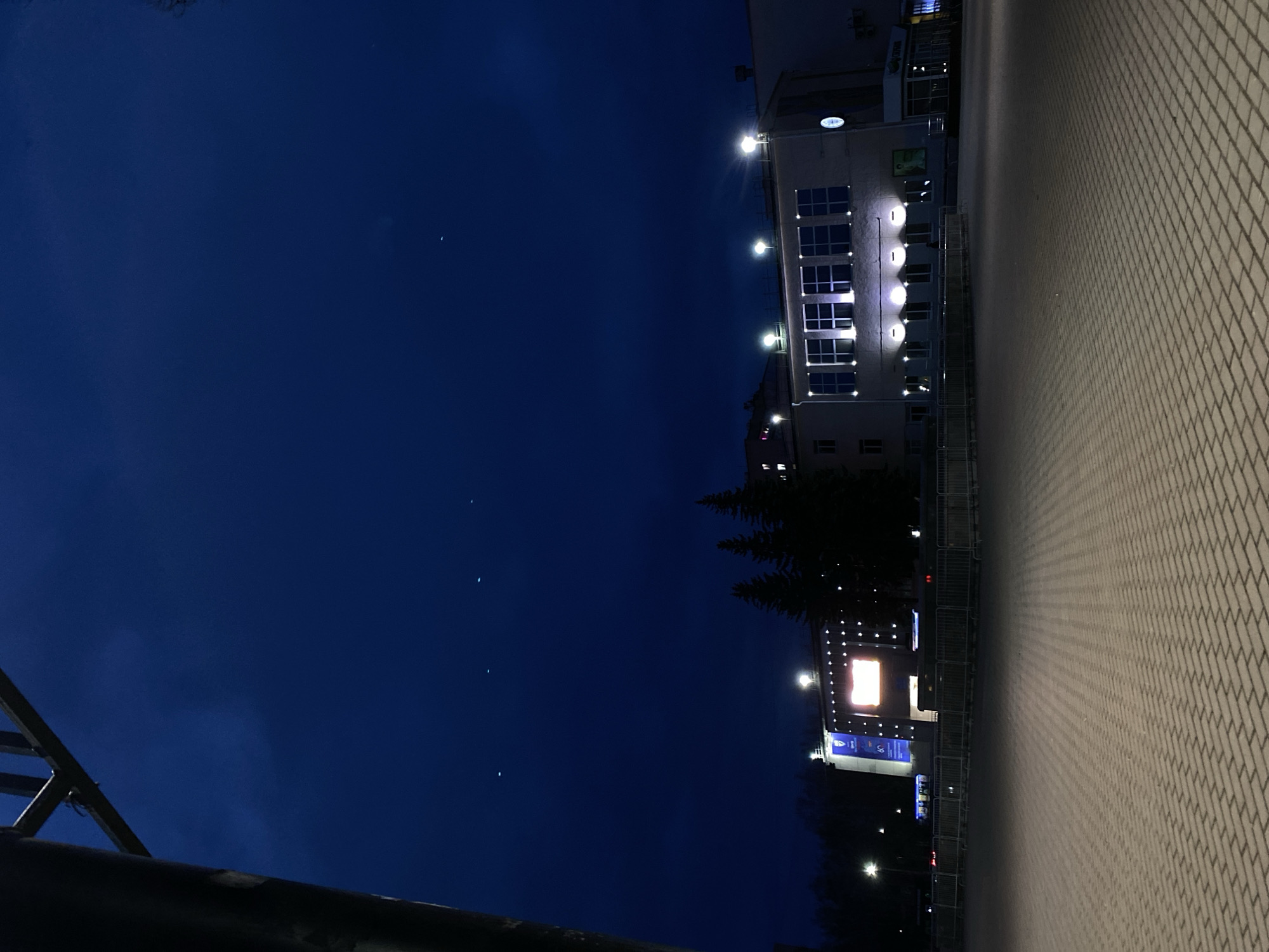 The image depicts an outdoor night scene in what appears to be a public or commercial area in Chaikovsky, Russia. The time of day is night, characterized by a deep indigo-blue sky containing several faint, pinprick lights that could be stars or distant artificial lights. The weather appears clear.
The scene is devoid of people, presenting a quiet, static environment. The foreground features an indistinct, dark structure, possibly a railing or part of a bridge, partially obscuring the lower left. The right side of the image is dominated by a large expanse of light-colored, interlocking paver stones, creating a textured ground surface. Long, dark shadows are cast across this paved area from the left, suggesting a low light source or objects outside the frame.
In the mid-ground and background, several buildings are visible, illuminated by bright artificial lights. A building on the left features multiple rectangular windows and is adorned with several wall-mounted spotlights that emit a bright, cool light. Above its windows, a light-colored sign with some barely discernible text, possibly "Coruna," is present. Further into the background and to the right, another structure, possibly part of a complex or event venue, showcases a large, brightly lit screen displaying a vibrant orange and yellow image. Below this screen, a blue banner with white text is visible, though the text itself is too small to read. Coniferous trees are silhouetted against the dark sky between the buildings, and metal barriers or fencing are also present in front of the background structure. The overall impression is one of an illuminated urban space after dark.