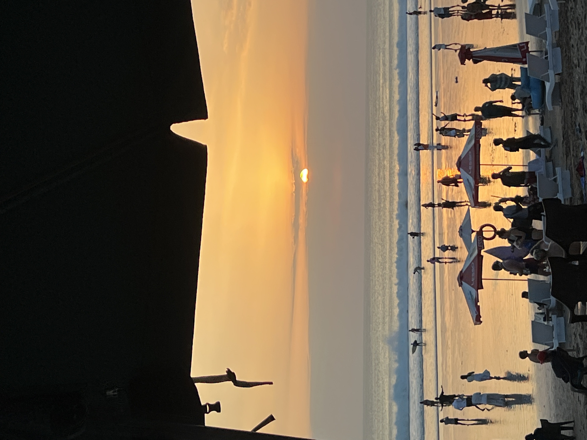 This is a vertical, eye-level, medium shot of a beach scene in Kuta, Indonesia, at sunset. The sun is setting over the ocean, casting a warm orange glow across the sky and reflecting on the wet sand. The waves are crashing on the shore, and several people are scattered across the beach. Some are walking, some are standing, and some are in the water. There are also a few umbrellas and beach chairs set up on the sand. The foreground is dominated by a dark, out-of-focus structure, likely an awning or canopy, that partially obscures the top of the image.