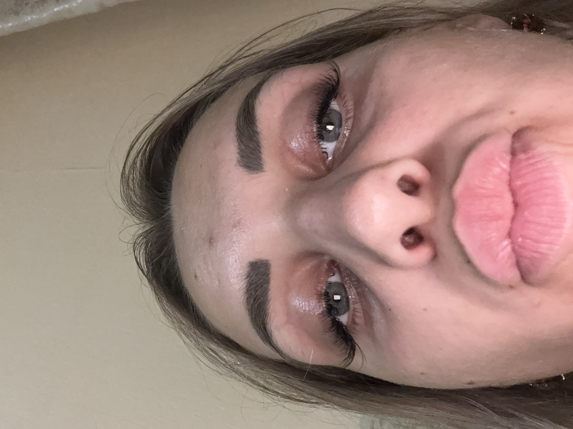 A close-up, low-angle shot shows a young woman's face, with her eyes and lips the primary focus. Her eyes are open, and she is wearing makeup, including mascara and false eyelashes. Her lips are pursed, and she appears to be wearing lipstick. Her eyebrows are well-defined. The background is a plain, light-colored wall. There is no indication of the city of Kholmsk, Russia, or any other specific location.
