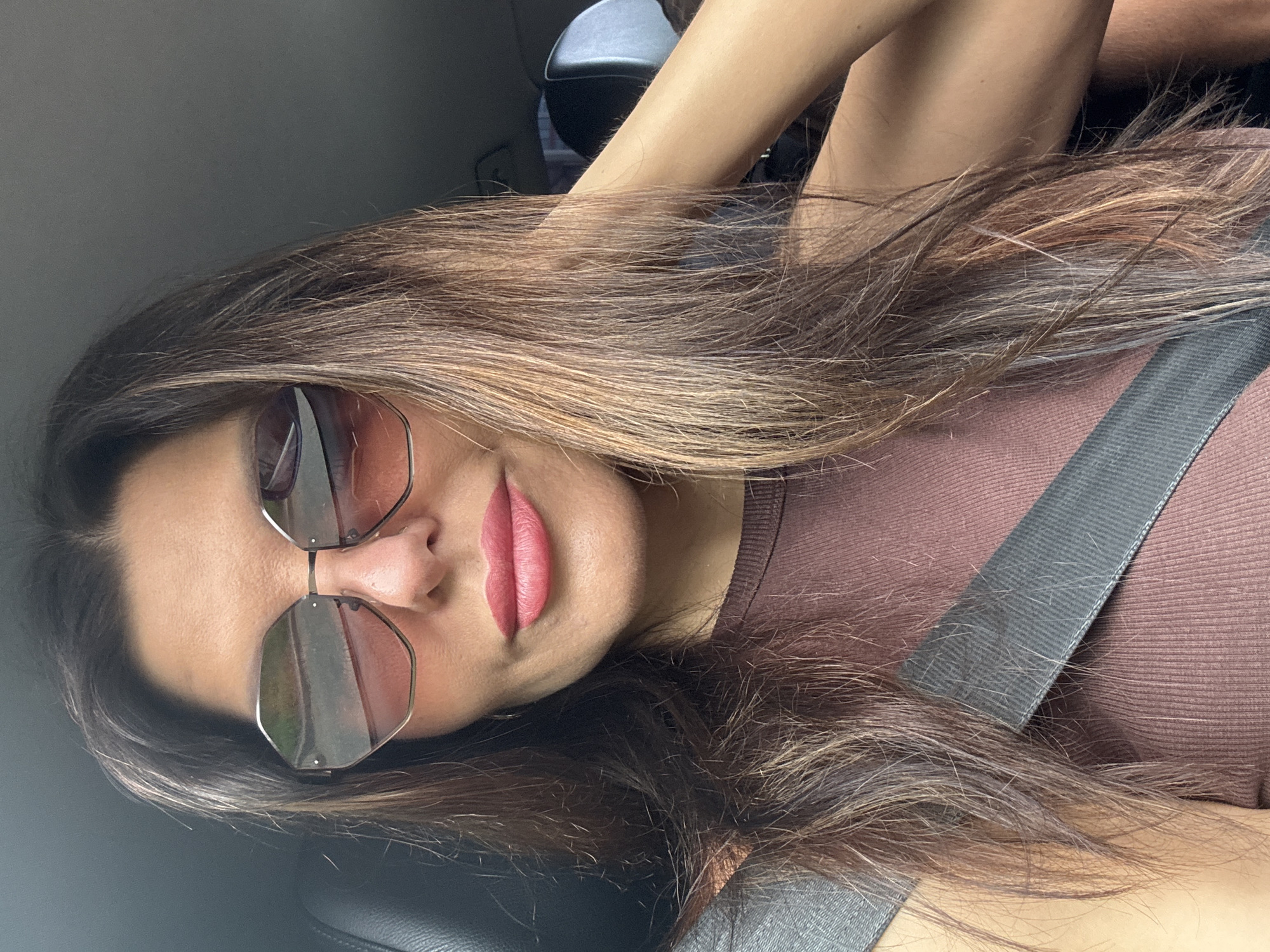 The image shows a woman in a car. She is wearing sunglasses and a brown top. Her hair is long and brown with highlights. A seatbelt is visible across her chest. The background is dark, suggesting the interior of a car. There is no indication of the location being Chaikovsky, Russia.