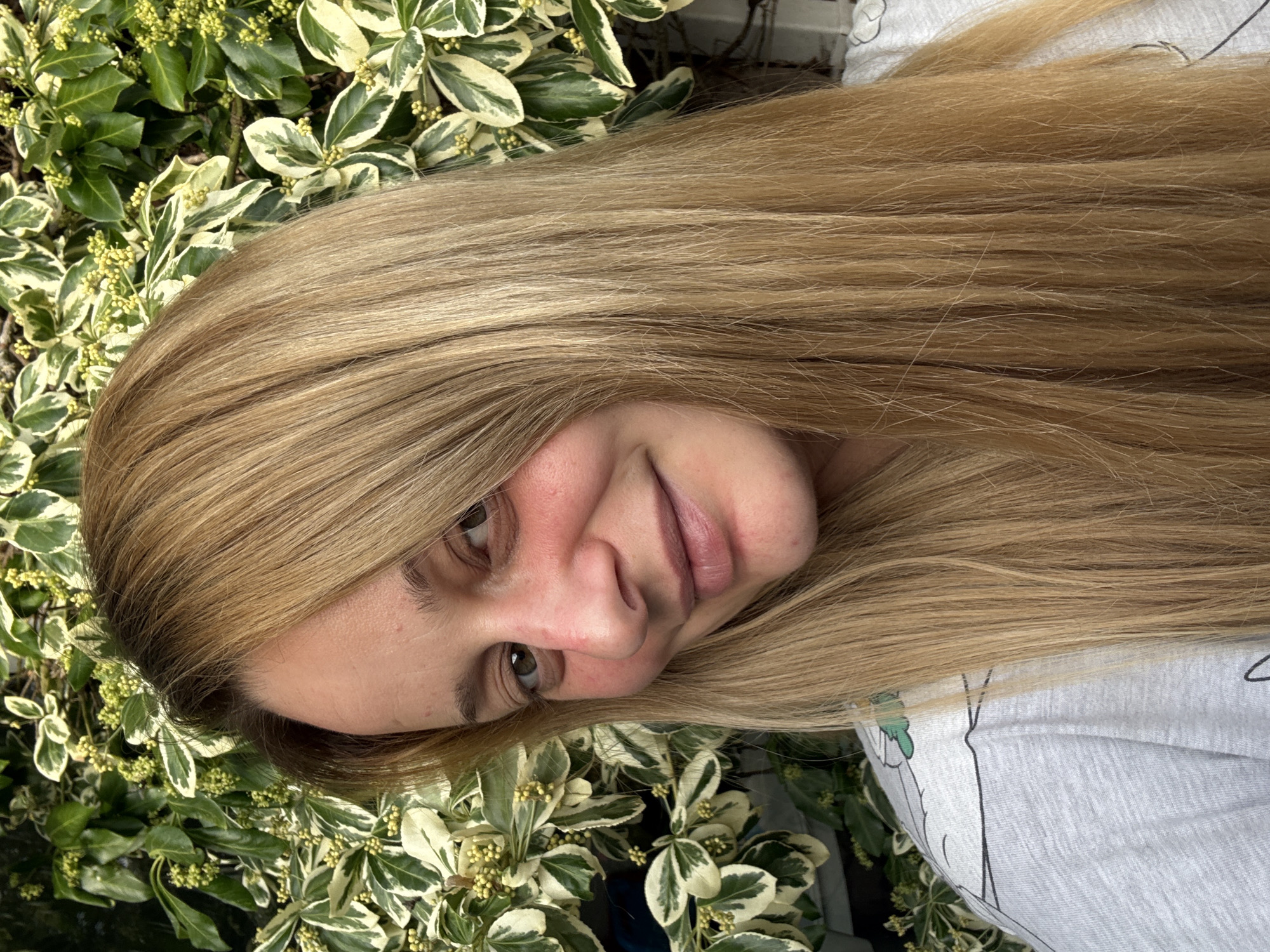 The image is a close-up portrait of a fair-skinned woman with long, blonde hair. She is positioned next to variegated green and white foliage. The woman has light brown eyes. She is wearing a light gray shirt with a graphic design on it. The background consists of more of the variegated foliage. The location is not discernible from the visual information provided.