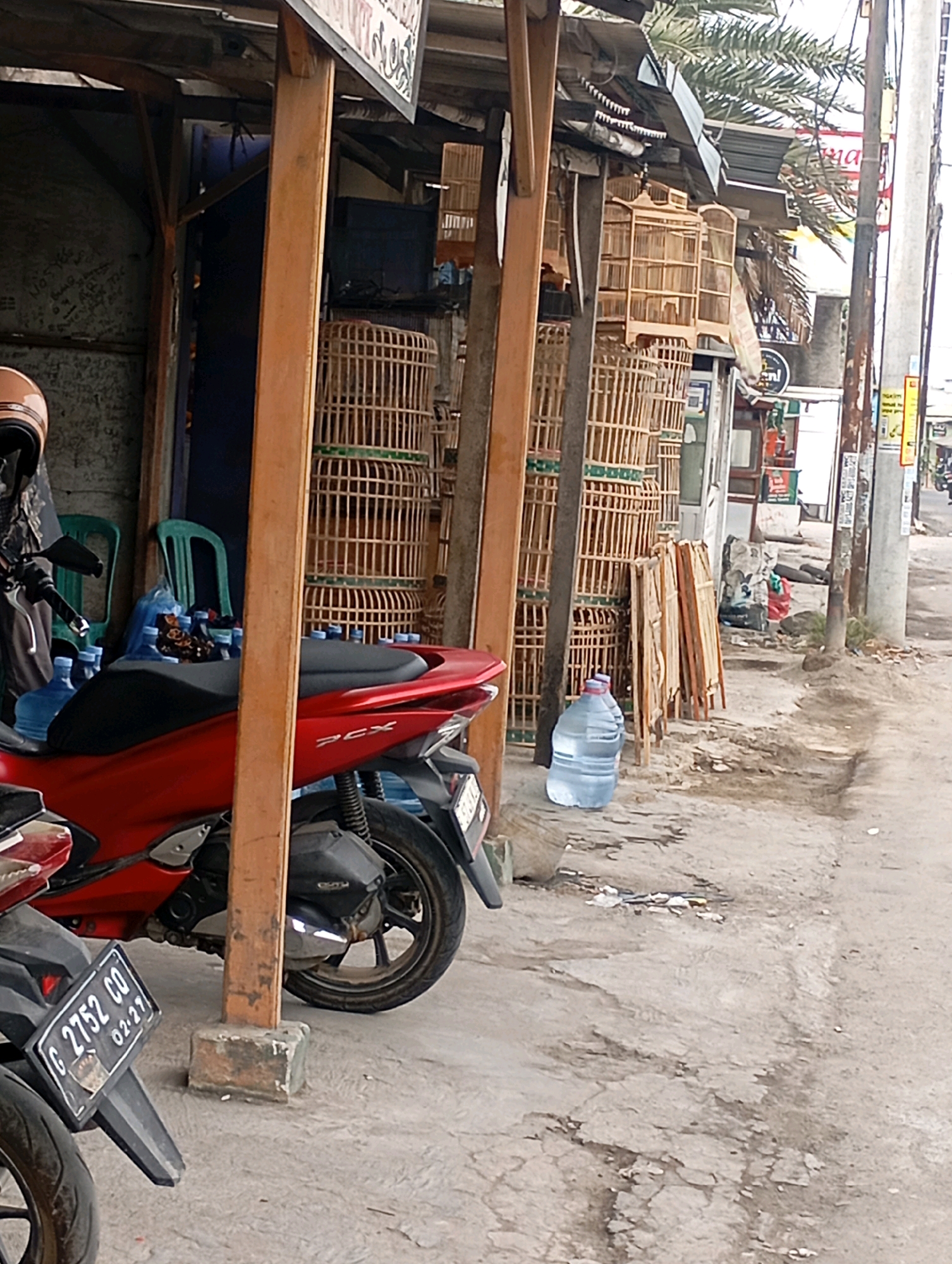 The image depicts a street scene in Babelan, Indonesia, likely a market or a roadside stall. In the foreground, a red motorcycle is parked, with another motorcycle partially visible. Several large blue water jugs are stacked near the motorcycles and in front of a makeshift stall. The stall is constructed with wooden posts and a corrugated metal roof, displaying numerous wooden bird cages of various sizes. Behind the stall, more bird cages are visible, along with some green plastic chairs. A palm tree is visible overhead, suggesting a tropical or subtropical environment. The ground is paved with concrete that appears worn and cracked in places, with some debris scattered around. Utility poles and wires are visible in the background, indicating an urban or semi-urban setting. The overall impression is that of a humble commercial area, possibly focused on selling birds or related items. There are no people clearly visible in the frame.