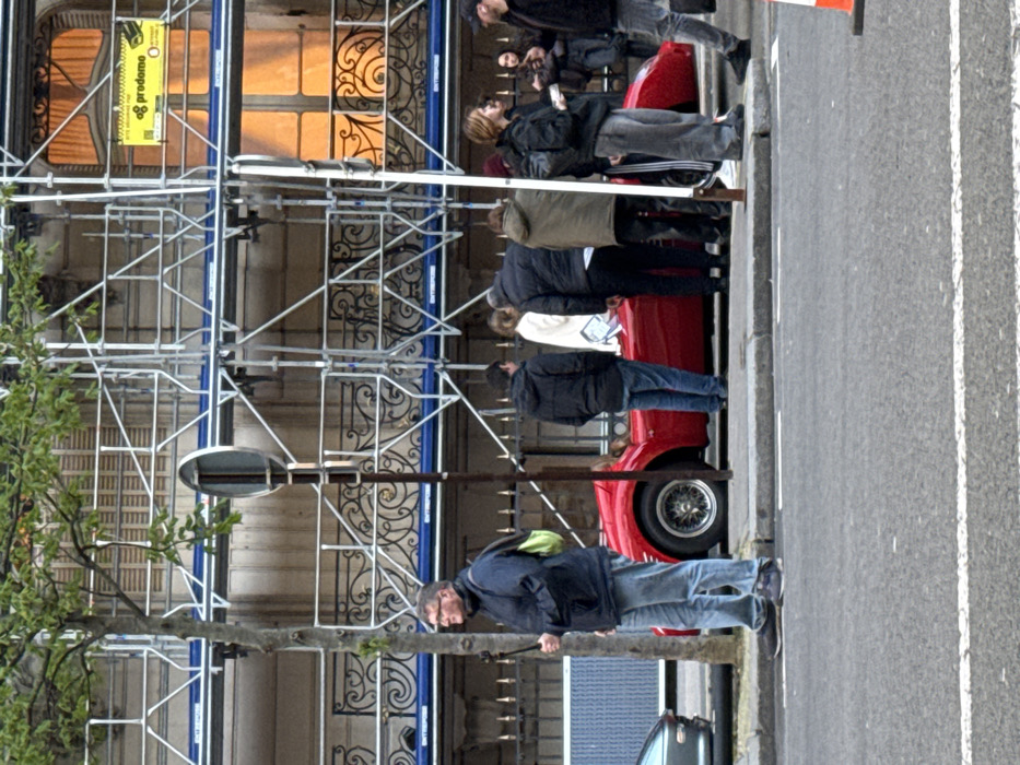 The image portrays an urban street scene in Paris, France. A multi-story building in the background, characterized by ornate iron balconies, is largely obscured by extensive silver metal scaffolding. Several upper-story windows exhibit an internal orange glow, and one window displays a yellow rectangular sign with "prodome" text.
At street level, a red two-seater sports car with wire wheels is parked at the curb, oriented towards the right. A group of approximately seven individuals, dressed in casual attire including various dark jackets and jeans, are clustered around the red vehicle, facing it. To the left of this group, a man wearing glasses, a dark jacket, blue jeans, and a lime green backpack stands on the sidewalk, holding a black selfie stick vertically. A young tree with sparse green leaves and a dark, circular street sign pole are positioned near this individual. The rear section of a silver vehicle is partially visible on the far left.
The foreground consists of an asphalt street with a white painted line and a concrete curb. The top of an orange traffic cone is discernible in the bottom right corner. The individuals appear to be observing or interacting within the vicinity of the red sports car.