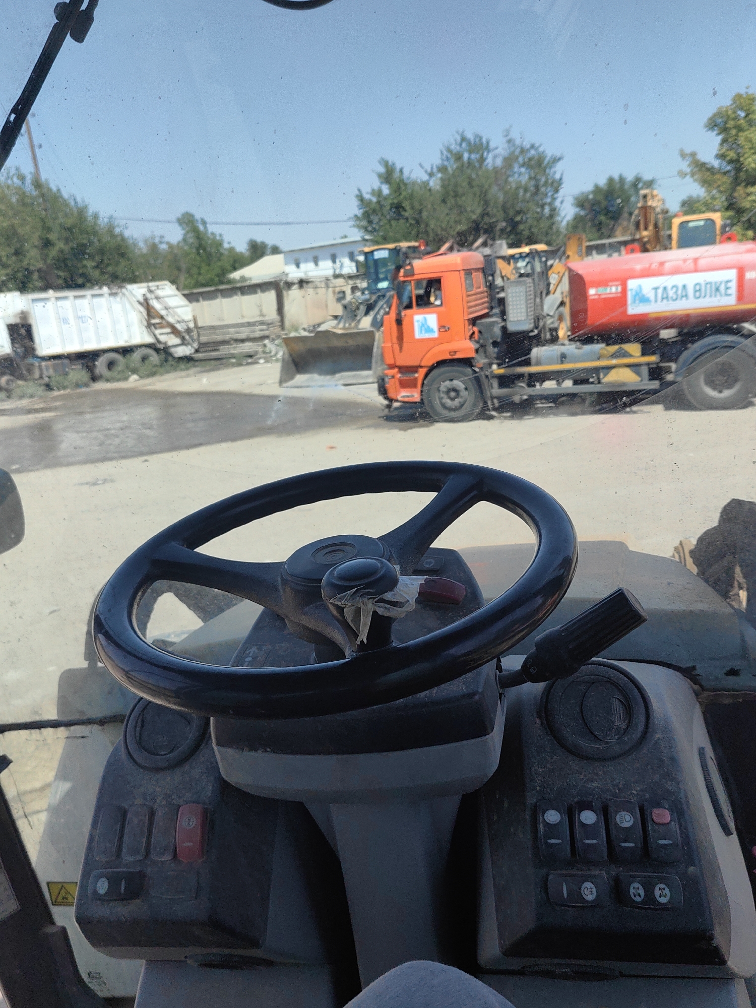 The image is a first-person view from inside a vehicle, likely a construction or heavy machinery operator's cabin. A black steering wheel is prominently displayed in the foreground, with a control lever to its right. To the left and right of the steering column are control panels with various switches and buttons, some of which appear dusty.
Outside the vehicle, a sunny outdoor scene is visible through the windshield. In the middle ground, an orange truck with a red tanker trailer is parked. The trailer has text in a Cyrillic script. Behind the orange truck, other heavy machinery, including excavators and a white garbage truck, are parked. The ground is dusty and appears to be a lot or yard. In the background, there are trees and some low-rise buildings. The sky is clear and blue. The scene suggests an industrial or storage area for vehicles, possibly a depot or recycling facility. The location is identified as Shymkent, Kazakhstan. There are no people clearly visible in the image.
