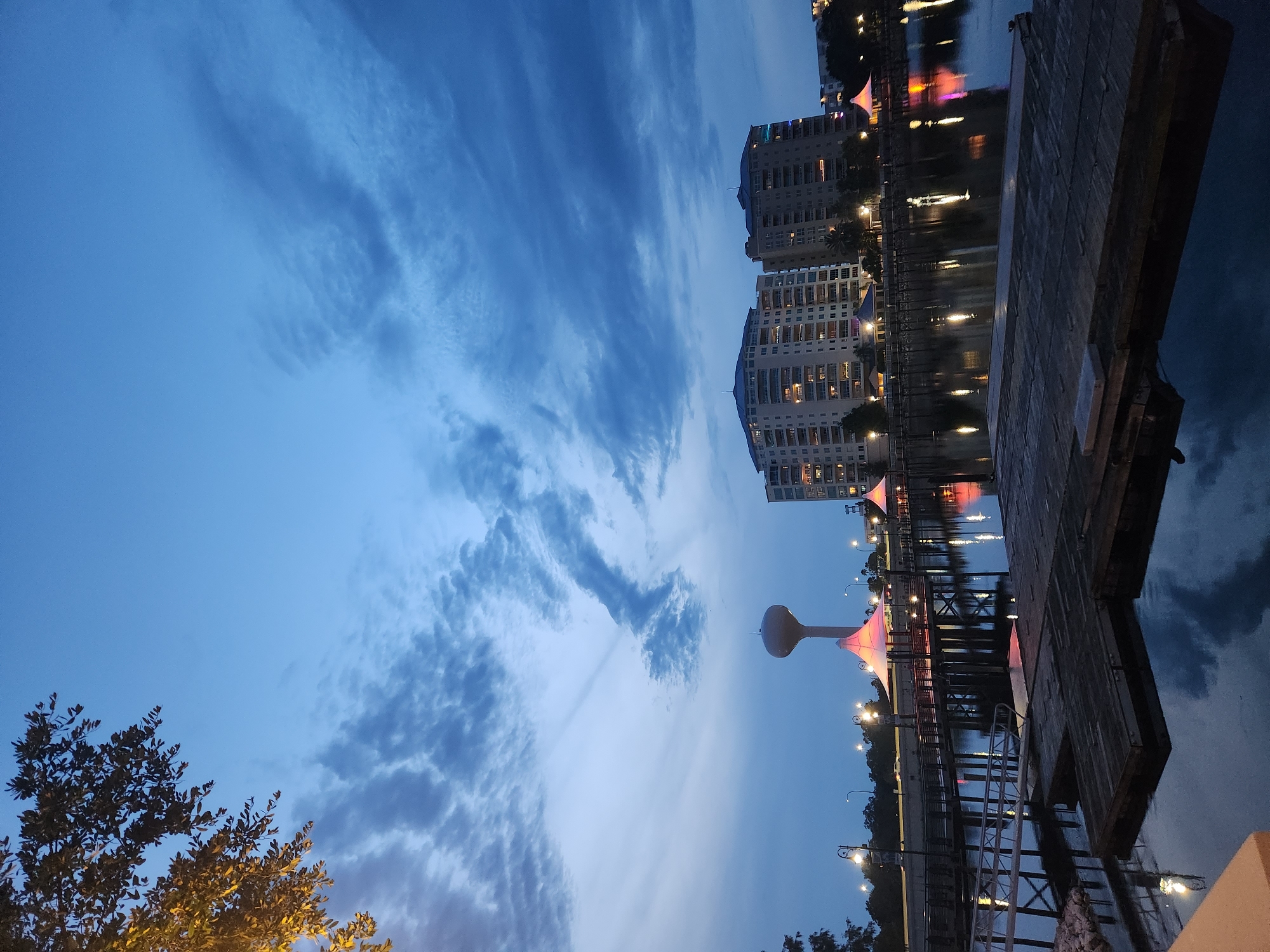 The image captures a twilight scene at Crane's Roost Lake in Altamonte Springs, Florida. The sky is a deep blue, transitioning from a lighter hue near the horizon, with scattered, wispy clouds. In the background, two large apartment buildings stand illuminated, their windows glowing with interior lights. To the left of the buildings, a distinctive water tower with a rounded top rises above the skyline.
In the middle ground, a boardwalk or pier extends over the dark water, lined with lampposts that cast reflections on the surface. Several brightly lit, tent-like structures with red and pink hues are positioned along the boardwalk, suggesting a public event or gathering space. In the foreground, a wooden dock or platform floats on the calm water, its rough texture and weathered appearance contrasting with the cleaner lines of the background structures. The water reflects the lights from the buildings, tents, and sky, creating a mirror-like effect. A large tree branch with green leaves hangs into the frame from the top left corner, partially illuminated by a warm light source. The overall atmosphere is serene and slightly moody, characteristic of dusk in a developed urban waterfront area.