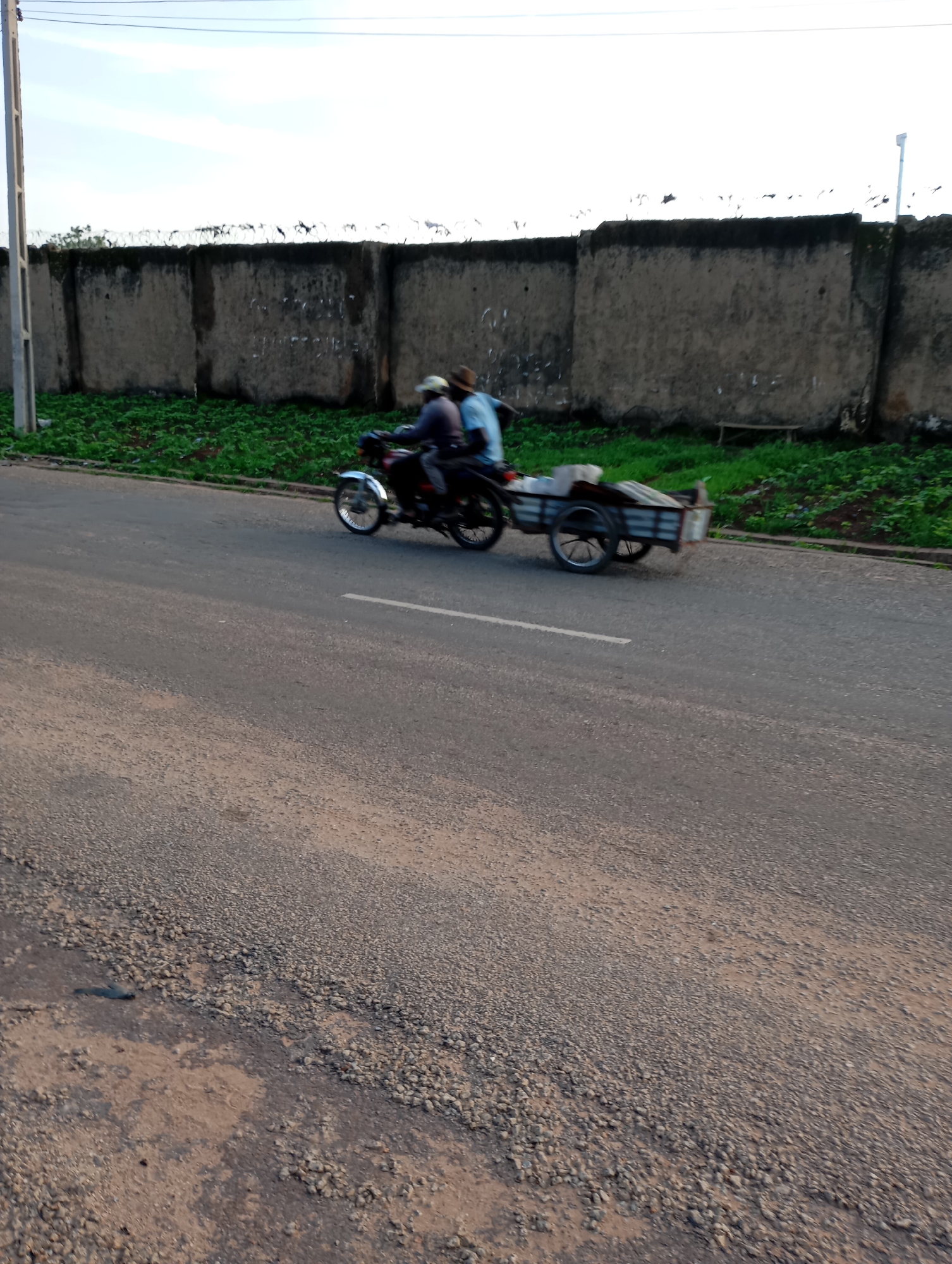 A motorcycle with two occupants and a trailer is traveling on a paved road in front of a concrete wall topped with barbed wire. The motorcycle, a dark color, has a rider wearing a hat and a passenger behind them wearing a light blue shirt. The trailer, attached to the rear of the motorcycle, is grey with two wheels and appears to be carrying some items covered with a tarp. The road has a white line marking. Green vegetation grows along the base of the wall. A light pole is visible on the left side of the image, and a lamp post is visible in the distance on the right. The sky is bright and overcast. The scene is outdoors, possibly in an urban or semi-urban area within Shendam, Nigeria.