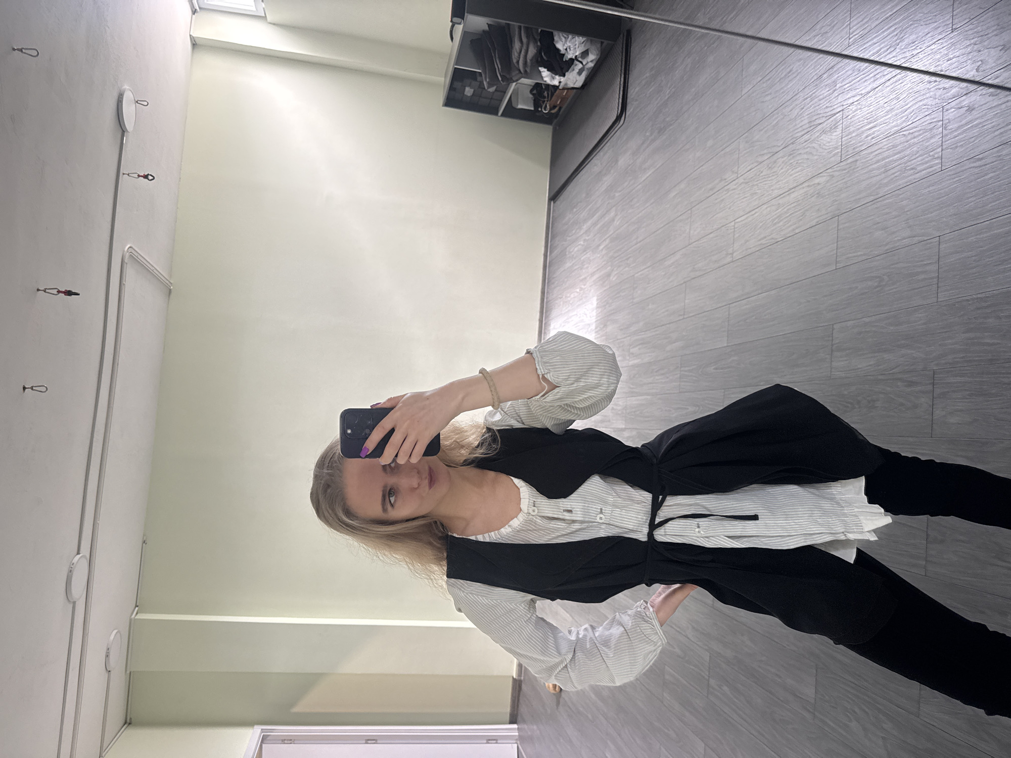 A young woman with blonde hair is seen taking a mirror selfie in an indoor setting. She is holding a dark-colored smartphone with her right hand, which has purple nail polish. She has a light-colored bracelet on her left wrist. Her attire consists of a long-sleeved, striped top in white and grey, featuring a scoop neckline with buttons. Over this, she wears a black sleeveless vest or tunic that ties at the waist with a black string. She is also wearing black pants.
The room, as reflected in the mirror, features light green or beige walls and a grey wood-plank patterned floor. On the wall behind her reflection, there are several silver hooks, some appearing to have red and black components attached, and white circular fixtures that could be lights or sensors. Cables or conduits run along the wall. In the upper right corner of the reflection, a black shelving unit or cabinet is visible, containing folded clothes or various items. A window or ventilation opening is present at the top of the wall. A metallic bar or rod also extends horizontally across the upper portion of the reflected floor area. The lighting in the room appears bright. No discernible text is visible in the image.