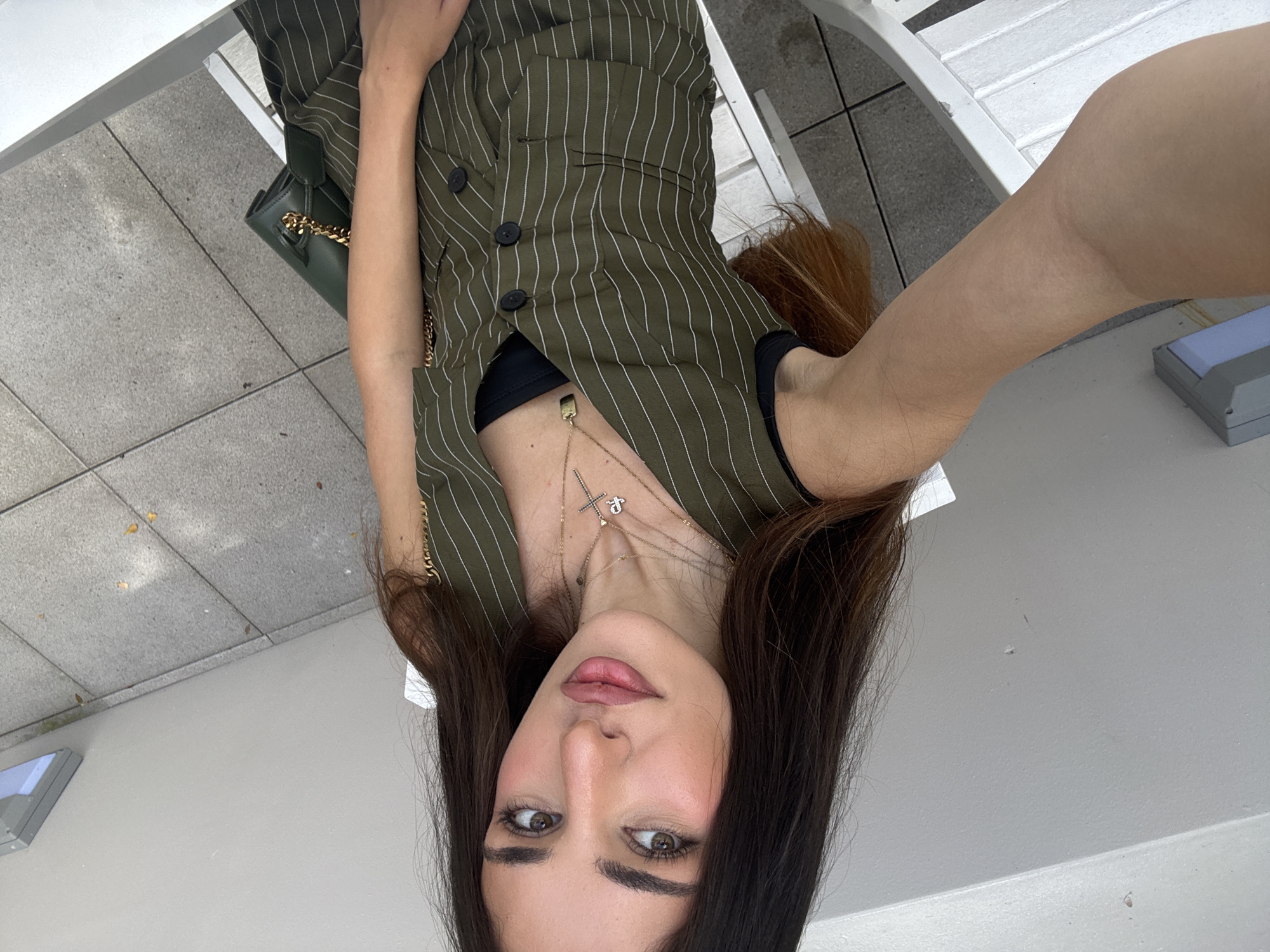 A young adult woman with long dark hair and light skin, wearing a green pinstripe vest over a black top and a green purse with a gold chain, is taking a selfie outdoors. She is sitting on a white chair on a patio with gray tiled flooring. The woman is wearing necklaces, one with a cross pendant and another with a rectangular pendant. The background includes a light gray wall and a light fixture mounted on it. The scene is set in Hallandale Beach, Florida, United States of America.