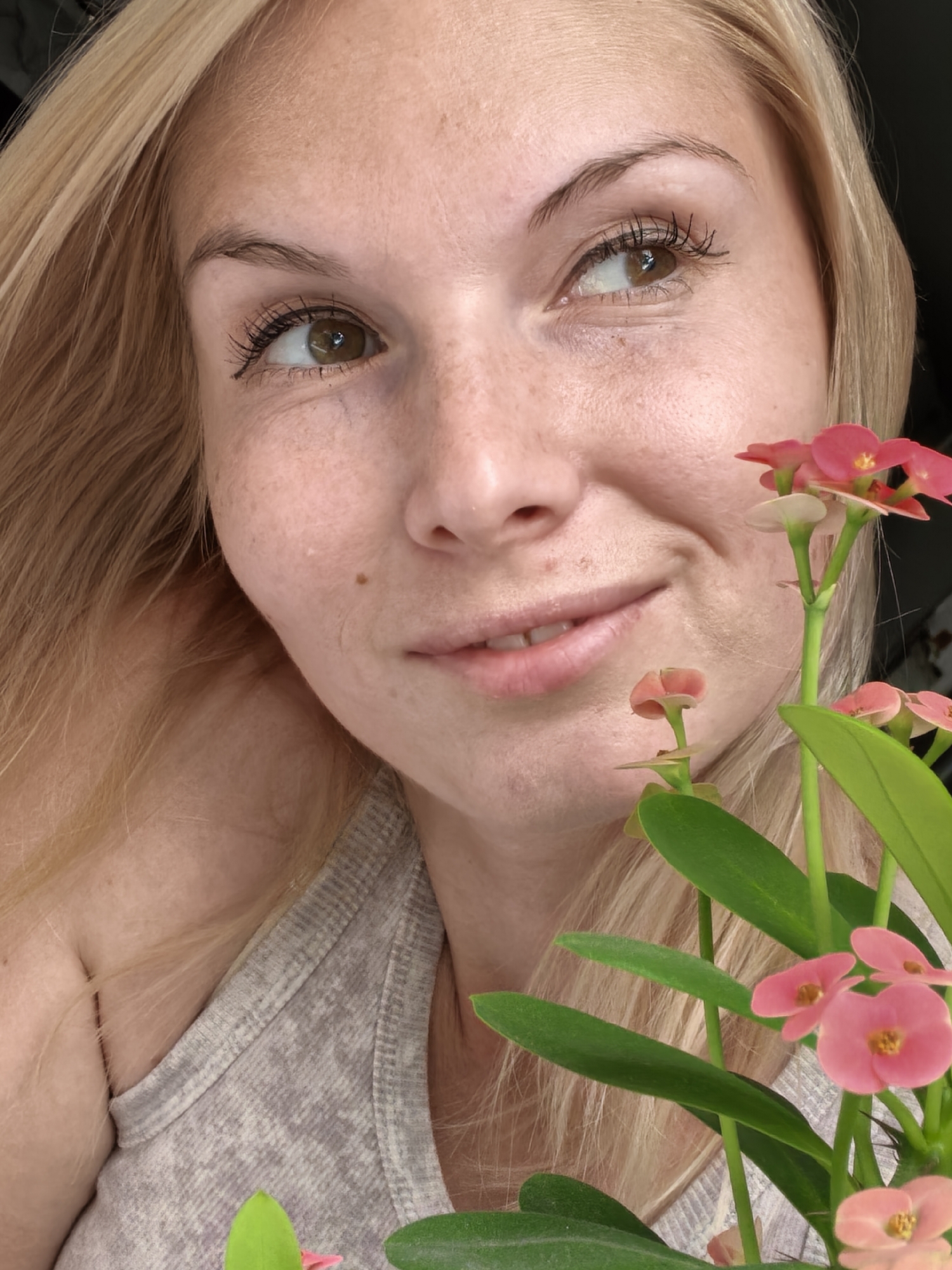The image is a close-up portrait of a woman with blonde hair and light skin with freckles. She is wearing a grey tank top and is looking upwards and to the right with a slight smile. In the foreground, to the right of the woman, is a plant with green leaves and small pink flowers. The background is dark and out of focus. The image appears to be taken indoors.