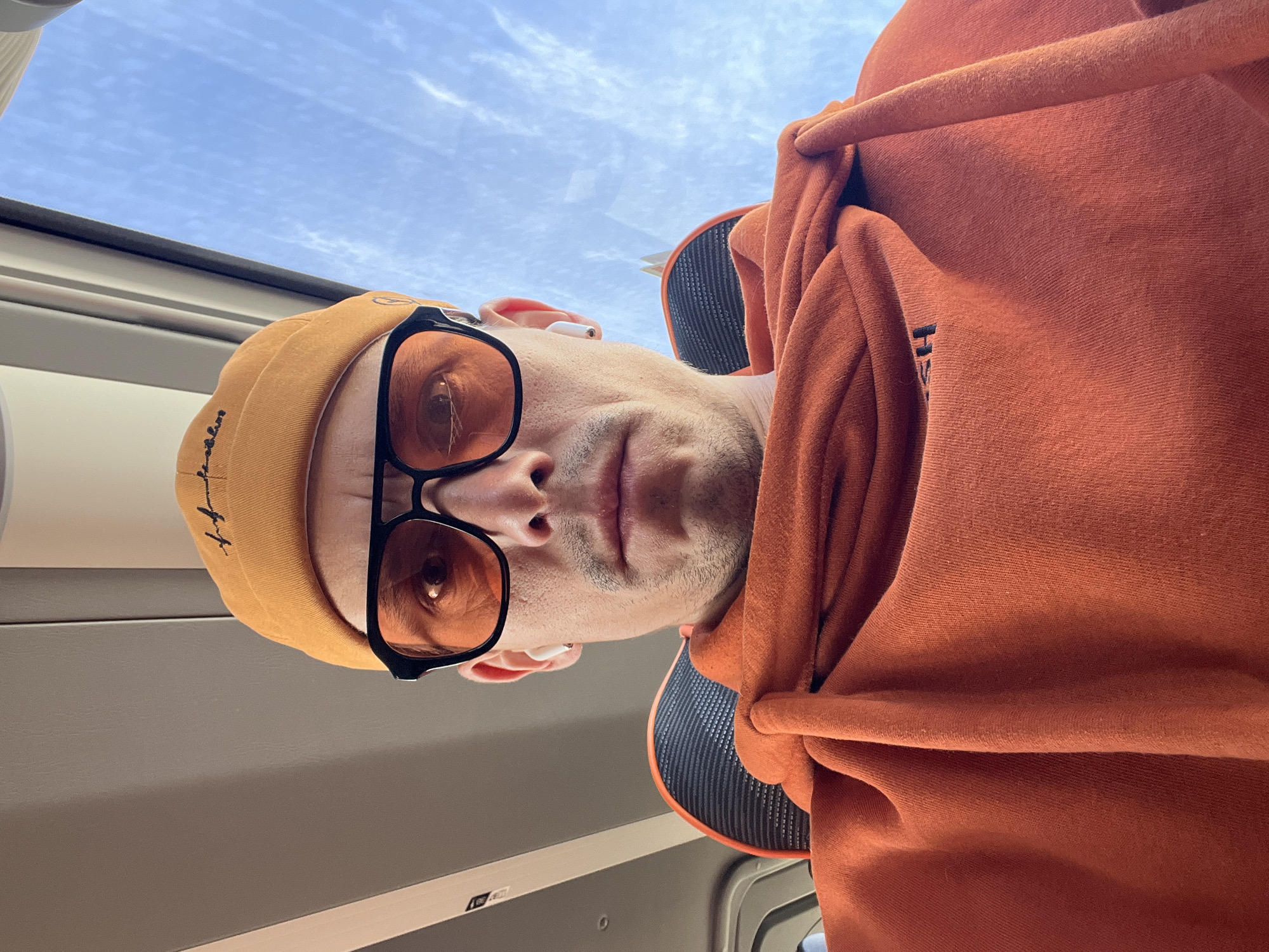 The image is a rotated, low-angle selfie of a man, largely occupying the central and left portions of the frame. He is looking directly at the camera with a neutral expression, wearing an orange beanie with faint black text or a logo, and large black-framed sunglasses with amber-tinted lenses. A white earbud is visible in his right ear. He has a slight stubble on his face and is dressed in a rust-orange hooded sweatshirt, with some black text, possibly an "H", visible on the shoulder area.
Behind his head, on the right side of the image, is a black and grey textured panel, likely a headrest or a part of the vehicle's interior. The upper left portion of the image reveals a bright blue sky with scattered white clouds, visible through a window, indicating a daytime setting. The lower section shows the light-colored interior lining and window frame of what appears to be a moving vehicle, such as a car or train. The overall impression is a casual, in-transit moment in Chaikovsky, Russia.