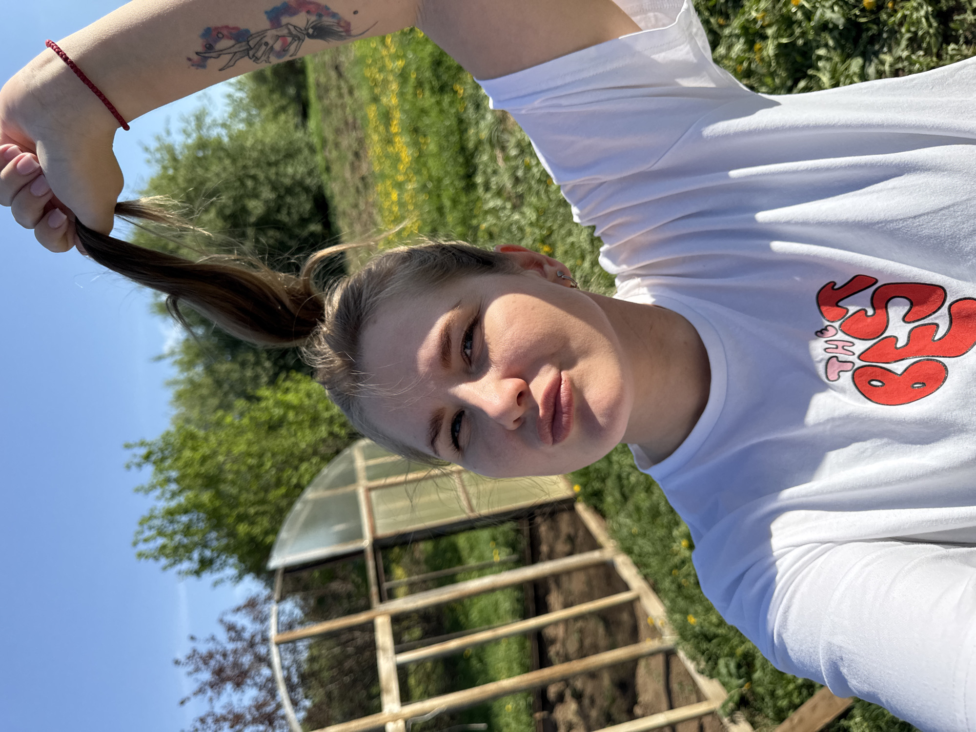 A woman with light skin and blonde hair pulled into a high ponytail stands outdoors near a greenhouse. She is holding the end of her ponytail. The woman is wearing a white t-shirt with a design on it and has a tattoo on her left forearm. In the background, a clear blue sky is visible over a landscape with green grass, trees, and dandelions. The location is Фоки, Russia.