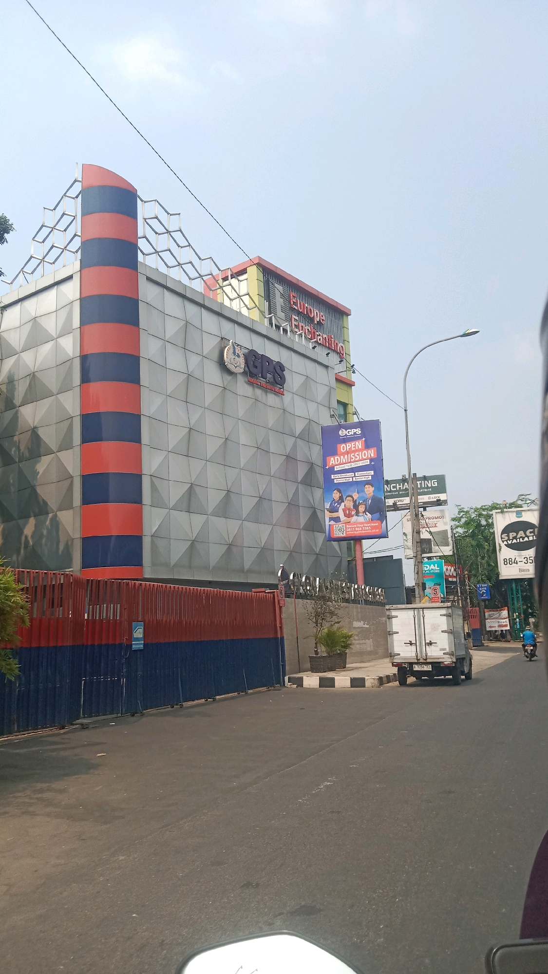 A low-angle, full-shot photograph, taken outdoors on a sunny day, shows the front of a modern, multi-story commercial building in Bekasi, Indonesia. The building has a grey, geometric facade with blue and orange striped accents on a vertical column on the left side. A large sign with "Europe Enchanting" is visible at the top of the building. To the right of the building, a large billboard advertises "GPS Open Admission" with images of children and adults. A lamppost stands in front of the billboard. The street in front of the building is paved with asphalt and has a sidewalk on the right. A white truck is parked on the side of the road. A person on a motorcycle is seen in the distance on the right side of the image. The sky is blue with some clouds. A red and blue fence encloses the property.