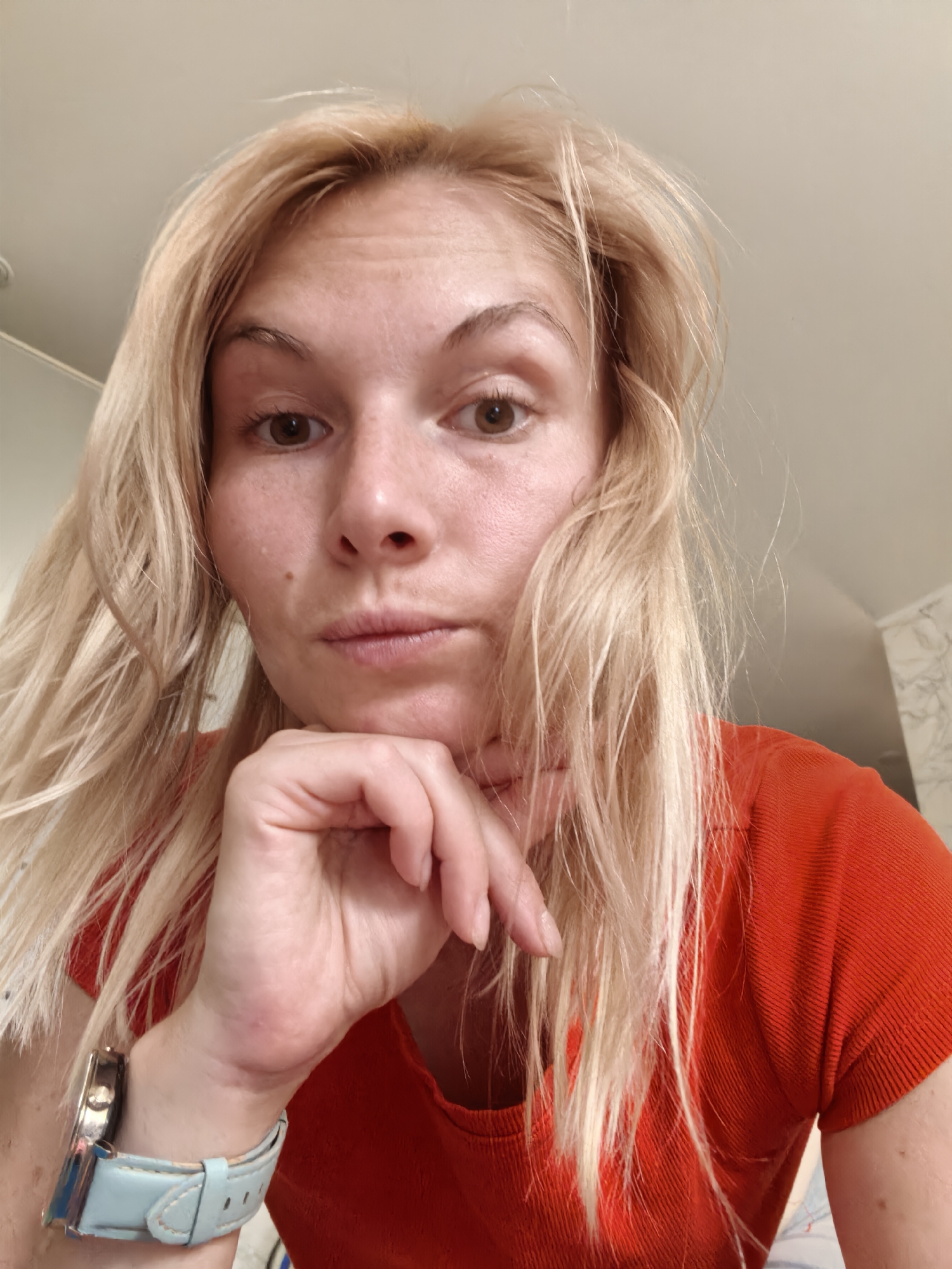 The image is a close-up selfie of a woman with blonde hair, wearing an orange t-shirt and a light blue watch. She is resting her chin on her hand and looking at the camera with a neutral expression. The background appears to be an indoor setting with neutral-colored walls and some blurry patterned wallpaper on the right side. There is no indication of the city of Sterlitamak, Russia in the image.