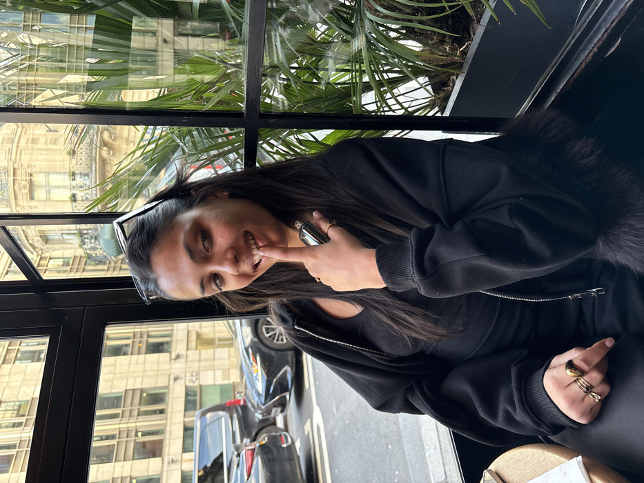 The image displays a head-and-torso shot of a woman seated indoors, looking out through a large window. The image is rotated 90 degrees counter-clockwise.
The woman is the central focus, positioned on the right side of the frame. She has long, dark hair, dark eyes, and is smiling broadly, revealing her upper teeth. She is wearing sunglasses pushed up onto her head. Her right index finger is pressed against her mouth, near her upper teeth, in a playful or thoughtful gesture. She wears multiple gold-toned rings on her right hand, including a distinctive stacked ring on her index finger and another on her ring finger. Her left hand holds a small, dark cylindrical object, possibly a vape or a small bottle, near her chest.
She is dressed in a black long-sleeved top or hoodie, which appears to have a zipper down the front, partially visible. A fuzzy, dark, fur-like material is visible around her right shoulder and upper arm, suggesting a fur-lined collar, hood, or trim on her garment.
Behind the woman, a large window with black frames separates her from the outside. Through the window, green foliage, possibly palm fronds, can be seen, either as part of an indoor plant display or growing just outside. Reflections in the window reveal an urban environment: light-colored, multi-story buildings with classic architectural details, and several vehicles, including cars and vans, on a street below. The reflections indicate bright daylight conditions.
The overall impression is one of a relaxed moment, with the woman expressing a cheerful emotion.