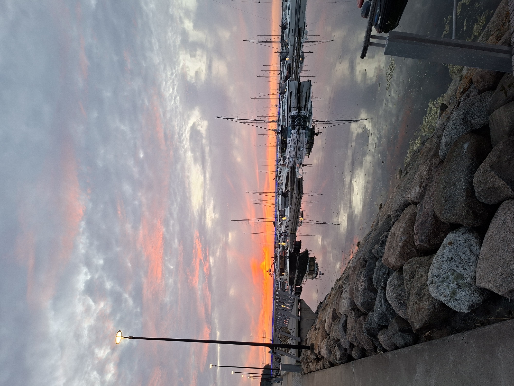 The image captures a serene and vibrant sunset or sunrise over a marina in Tallinn, Estonia. The sky is a dominant feature, filled with clouds illuminated by brilliant orange and red light near the horizon, transitioning to soft blues and grays higher up. This dramatic coloration is beautifully mirrored on the calm water surface, which also reflects the silhouettes of numerous boats.
A long line of yachts and sailboats is docked along a pier or jetty, their masts and rigging creating intricate patterns against the colorful sky. The water around the boats appears slightly murky with some organic matter, possibly algae. To the right, a rugged breakwater constructed from large rocks extends into the distance, separating the water from the land.
In the foreground, a paved promenade or walkway runs parallel to the water's edge. Several streetlights are visible along this path, one prominently in the mid-left, glowing with a warm yellow light, confirming the low light conditions of either dusk or dawn. The overall atmosphere is peaceful and picturesque, highlighting the natural beauty of the coastal setting. No discernible text is visible in the image.