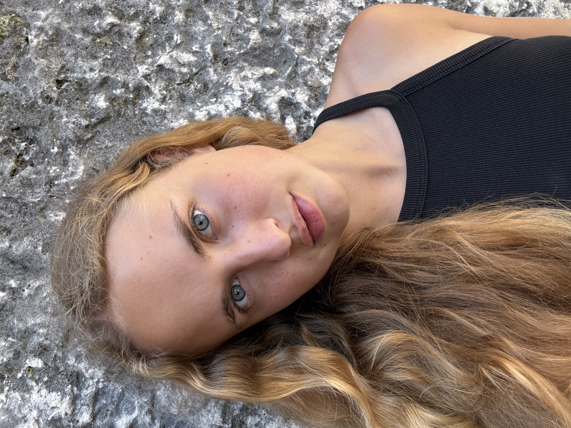 A young woman with long blonde hair is captured in a close-up portrait against a rough, grey stone wall. She is wearing a black ribbed tank top and her head is tilted to her left. Her blue eyes are open and looking towards the camera. The background is a textured stone surface, suggesting an outdoor environment. The lighting is natural and appears to be daylight.