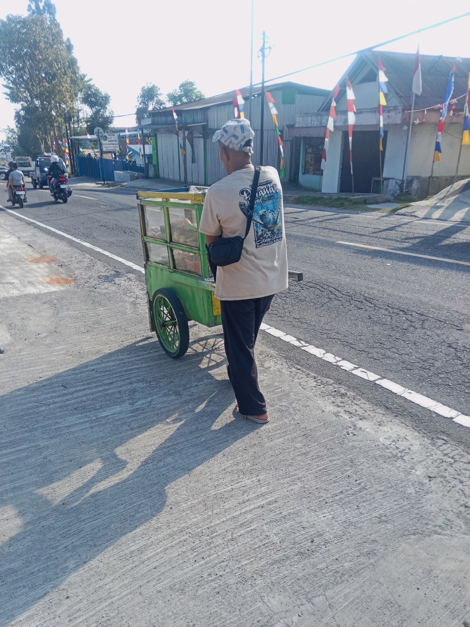 An outdoor, medium shot captures a man walking on the side of a paved road in Kertek, Indonesia. The man is viewed from behind, wearing a light-colored t-shirt with a graphic print, black pants, a patterned cap, and a dark bag slung across his chest. He is pushing a bright green food cart with a yellow trim. The cart has two wheels and glass panels, revealing food items inside. The road is asphalt with white markings, and the man is walking on a concrete sidewalk. To the left of the road, several motorcycles are visible, one with a passenger. In the background, buildings with corrugated metal roofs and walls are present, decorated with numerous Indonesian flags. Tall trees line the left side of the road, and the sky is bright and clear, indicating it is daytime. The sun casts long shadows on the sidewalk.