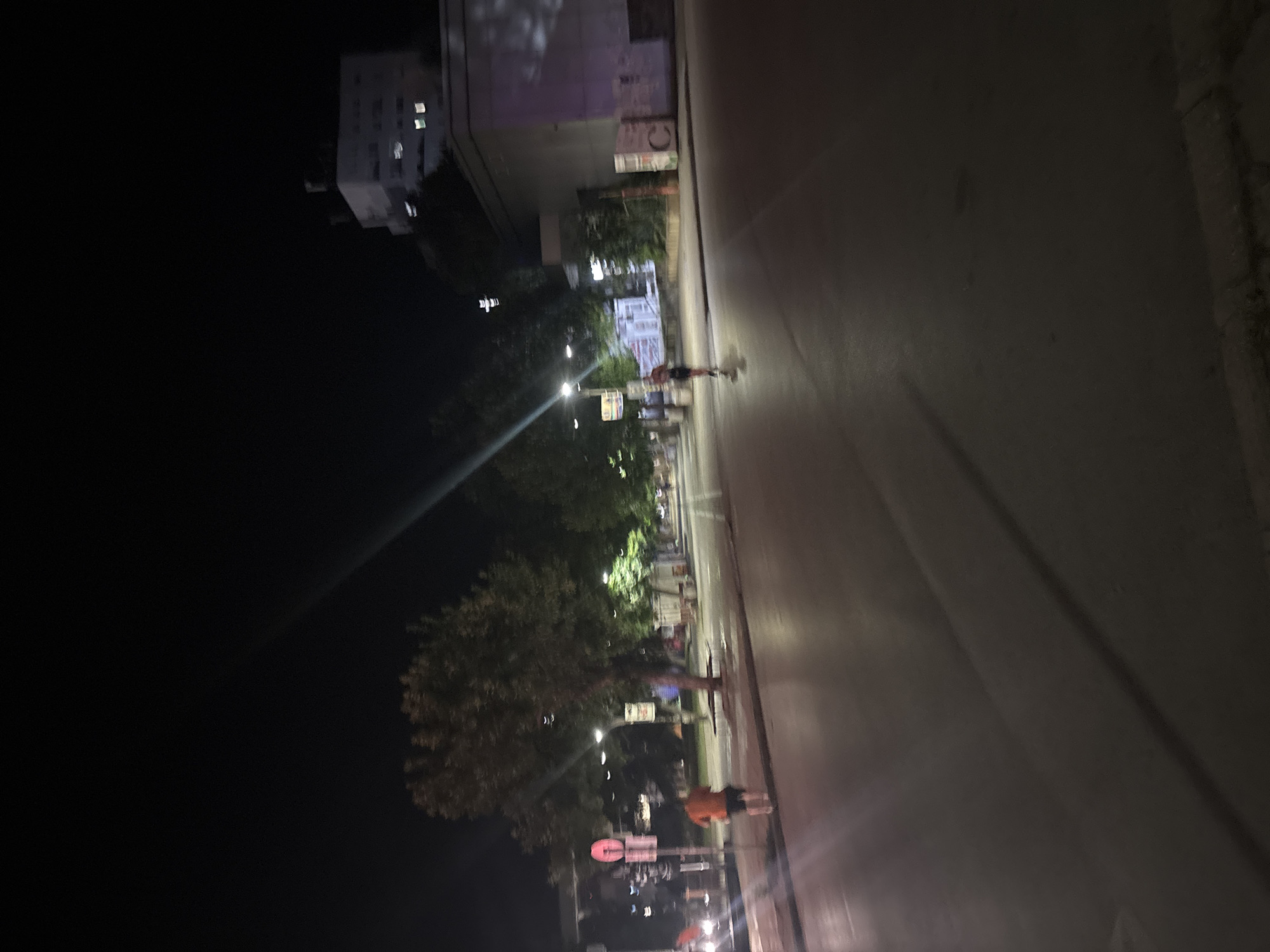 A nighttime outdoor shot shows a street with a person walking on the sidewalk and another person riding a scooter on the road. Trees line the sidewalk, and streetlights illuminate the scene. In the background, a building is visible with some windows lit. The road appears to be asphalt.