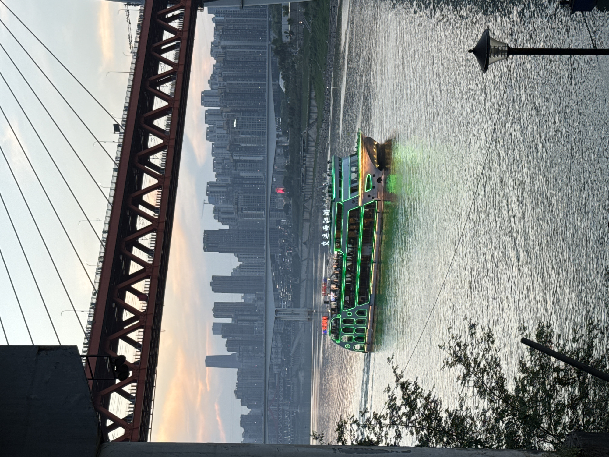 An evening cityscape view in Yuzhong District, Chongqing, China, shows a river with a brightly lit tour boat navigating the waters. The boat features green LED lights around its perimeter. On the left, the red, steel structure of the Qiansimen Bridge is visible. Across the river, there's a skyline of high-rise apartment buildings. The water's surface reflects the light. A street lamp is positioned on the right side of the frame, partially obscured by foliage in the foreground.