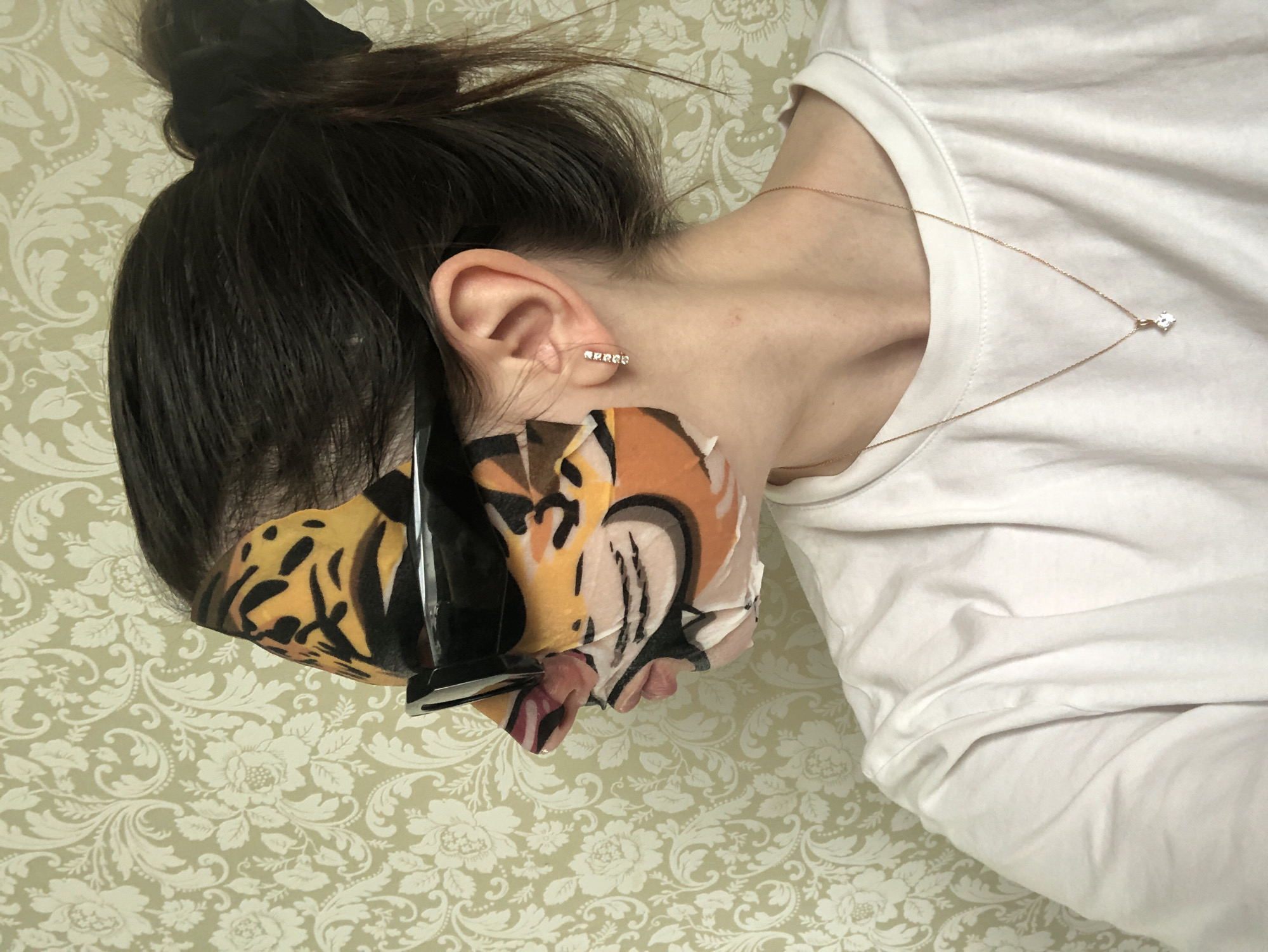 A woman with dark hair pulled up in a bun wears a tiger-striped facial mask and a white t-shirt. She is also wearing a gold necklace with a single pendant and a diamond stud earring. The background is a floral patterned wallpaper. The location is not determinable based on the image.