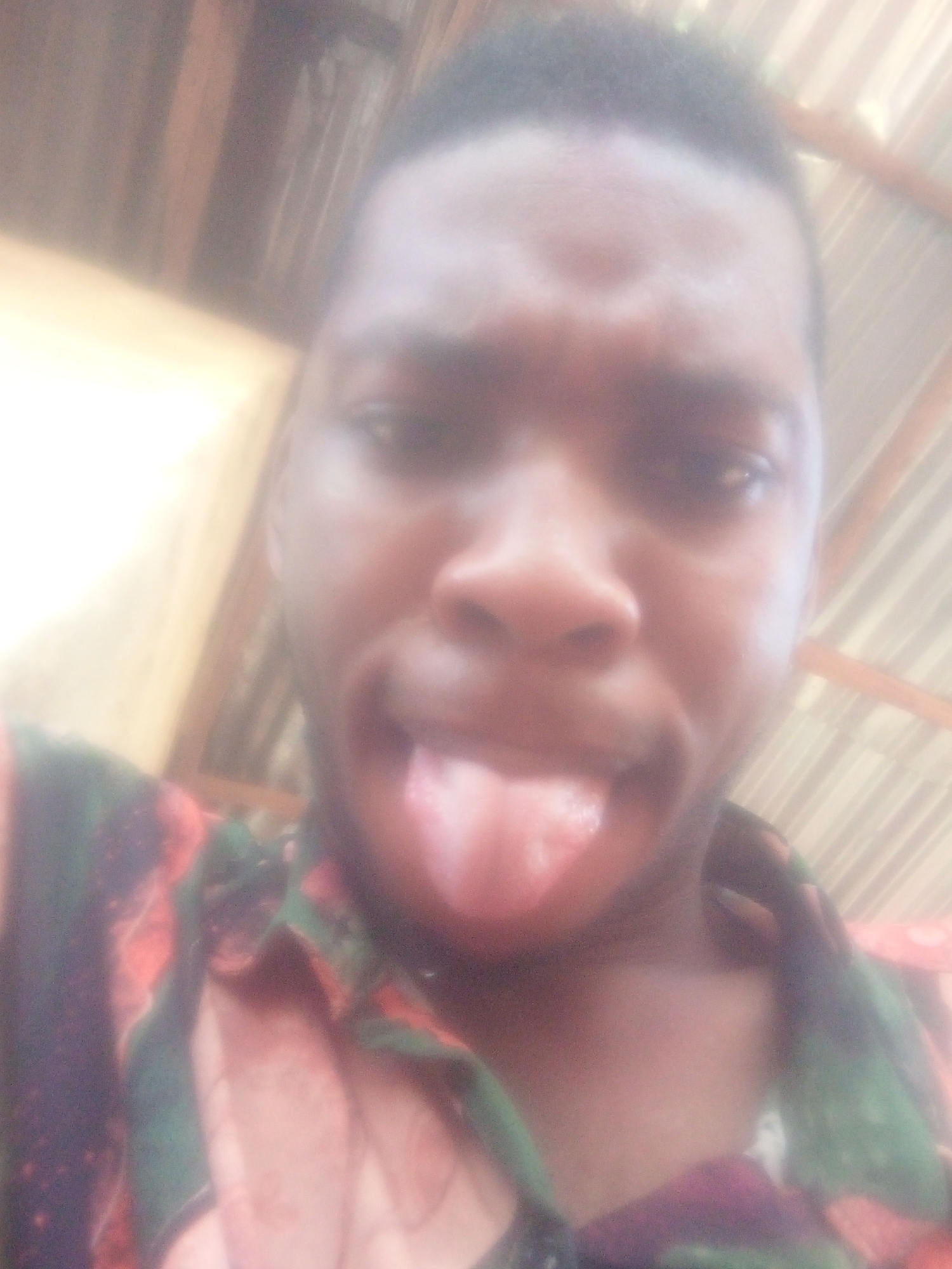 A close-up, slightly blurry selfie features a Black man sticking his tongue out. He wears a patterned shirt with green, red, and black hues. The background shows what appears to be part of a corrugated metal structure and a yellowish wall. The location is Unknown, Unknown.