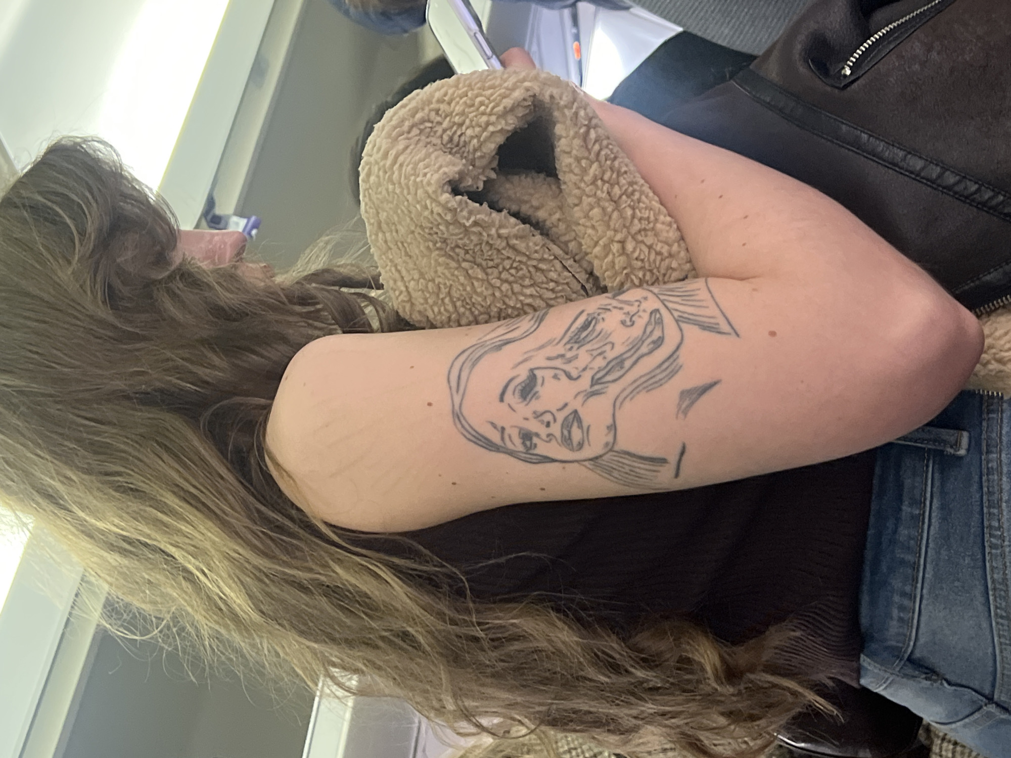 A woman with long brown hair is sitting on an airplane. She is wearing a brown sweater and a blue denim jacket. On her arm, there is a tattoo of a woman's face. She is holding a beige sherpa-lined jacket, with a dark brown leather jacket behind it. The location is an airplane cabin. There is no evidence to suggest that this image was taken in the city of Saint Petersburg, Russia.