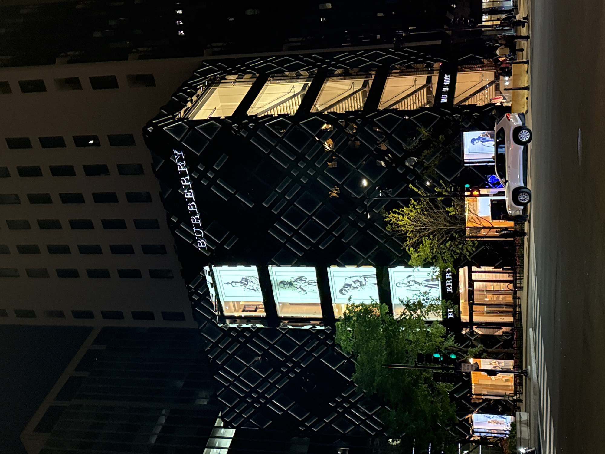 At night in Chicago, USA, the Burberry store stands prominently. The store's façade is a modern grid pattern with "BURBERRY" vertically displayed. The interior displays large monochrome images. A silver car is parked on the street near the store. Pedestrians are visible in the background. Streetlights illuminate the scene, casting reflections on the wet pavement. A neighboring building, with illuminated windows, is adjacent to the store. Lush green trees flank the Burberry store.