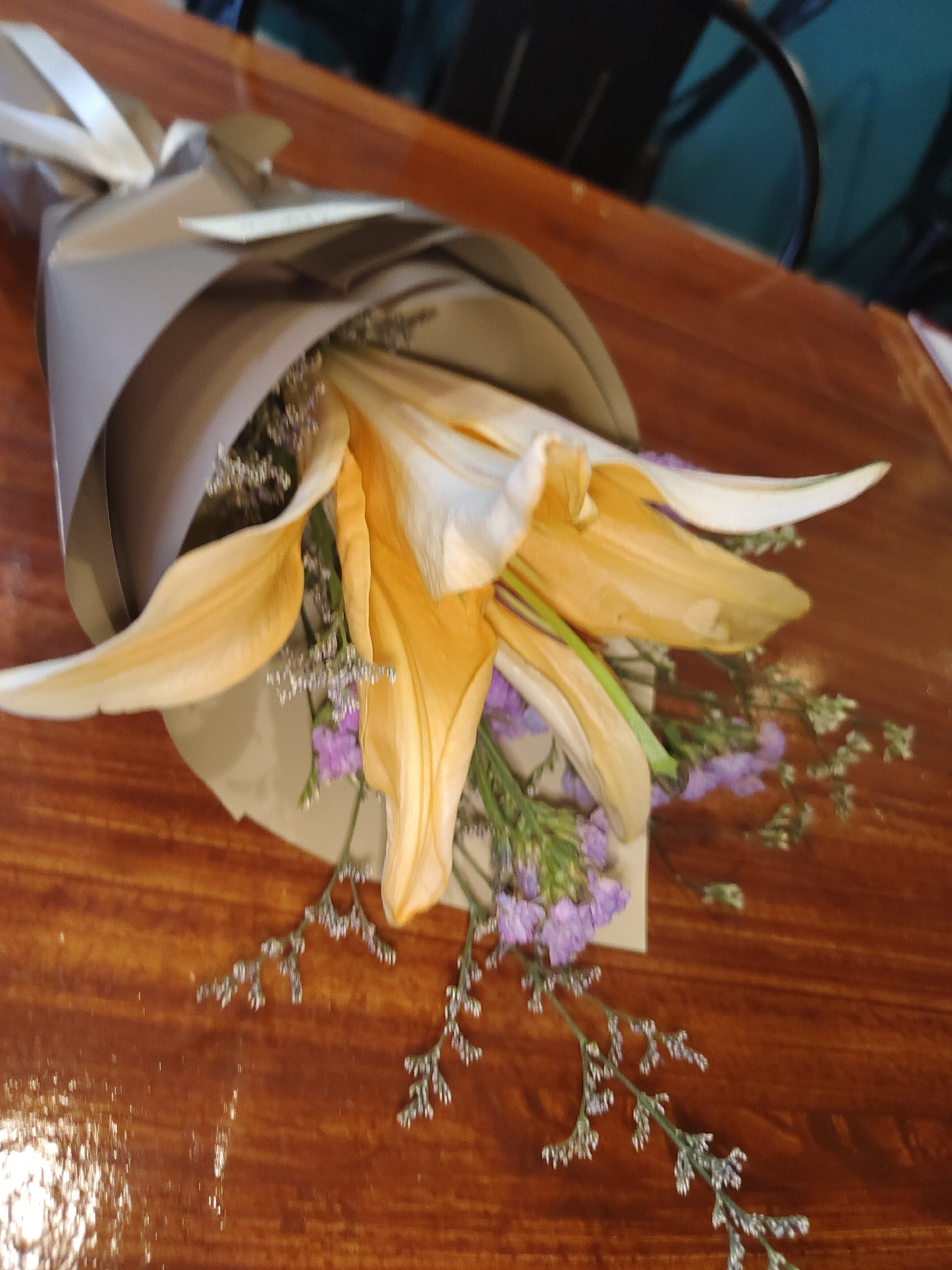A bouquet of light orange lilies and small purple flowers, wrapped in grey paper and tied with a white ribbon, rests on a polished wooden table. The table surface reflects light, creating a glossy appearance. The background is blurred, showing dark shapes that suggest furniture or possibly chairs. The scene appears to be indoors, in a setting that could be a cafe, restaurant, or a home dining area. There are no people visible in the image. The location is specified as Davao City, Philippines, however, there are no discernible landmarks or elements within the image that definitively place it in Davao City.