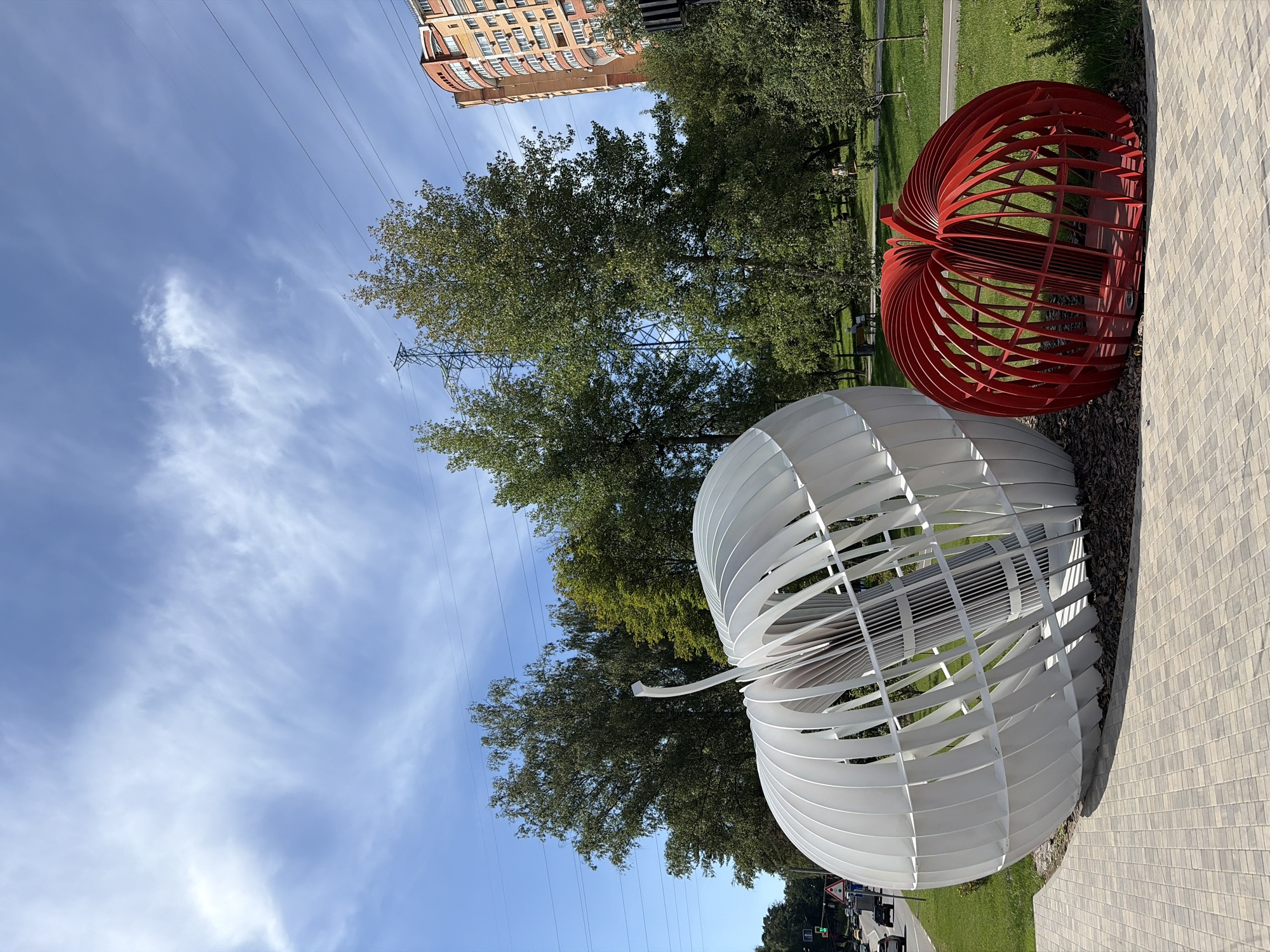 This is a photograph of a public art installation featuring two apple-shaped sculptures, one large white and one smaller red one, in Khimki, Russia. The sculptures are made of segmented metal frames and are placed on a paved area with a grassy park behind them. In the background, there are trees, a tall residential building with a curved facade, power lines, and a high-voltage electricity pylon. A street with cars and traffic lights is visible on the left side of the image. The sky is blue with scattered white clouds. There are a few people in the background of the park, sitting on benches and walking.