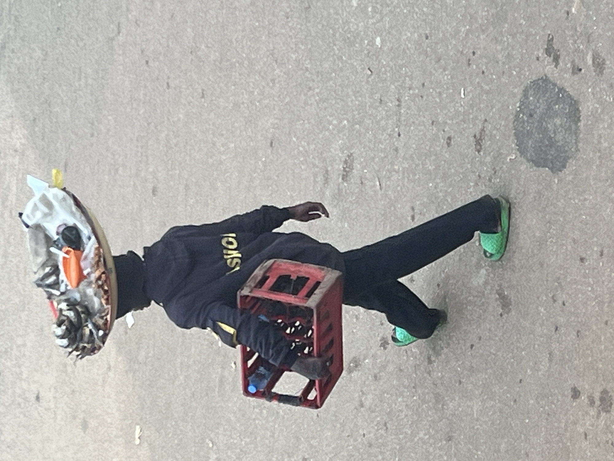 The image, taken in Bukuru, Nigeria, shows a person standing on a concrete surface. The person is wearing a dark hooded sweatshirt with yellow lettering on the sleeve that appears to read "IOIS" in reverse. They are also wearing black pants and bright green sandals.
The person is holding a red plastic crate filled with bottles. One blue bottle is visible. On their head, they are wearing a makeshift mask or head covering adorned with various items, including what looks like plastic wrap, orange material, and possibly food scraps or debris.
The scene appears to be outdoors, with the ground suggesting a street or walkway. The lighting indicates it could be daytime, but no specific time is evident. The overall impression is of a person engaged in collecting or transporting items, possibly for recycling or resale, given the debris on the headwear and the nature of the items in the crate. No discernible emotion is visible due to the obscured face.