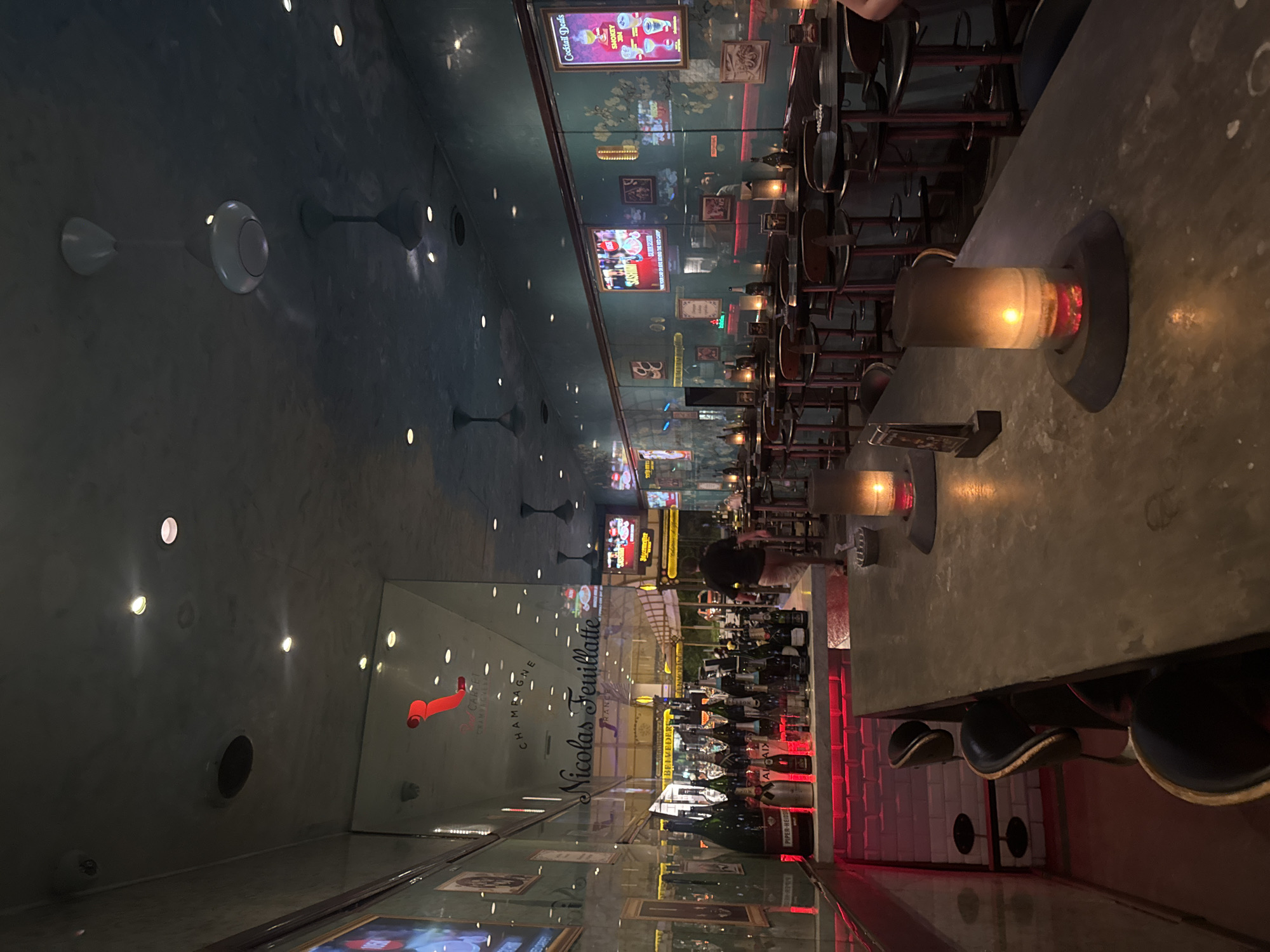 The image captures the interior of the "Red Carpet Champagne Bar" in Kuta, Indonesia, during nighttime. The bar features a long counter with several stools, and a well-stocked display of champagne bottles behind a glass partition. Red ambient lighting illuminates the lower section of the bar area, contrasting with the darker tones of the ceiling and walls. Multiple ceiling fans are visible, indicating a tropical climate. Digital screens displaying advertisements and entertainment are mounted on the wall. A single person is seen walking away from the camera towards the back of the bar. Several candle holders with lit candles are placed on the counter, adding to the intimate atmosphere of the establishment.