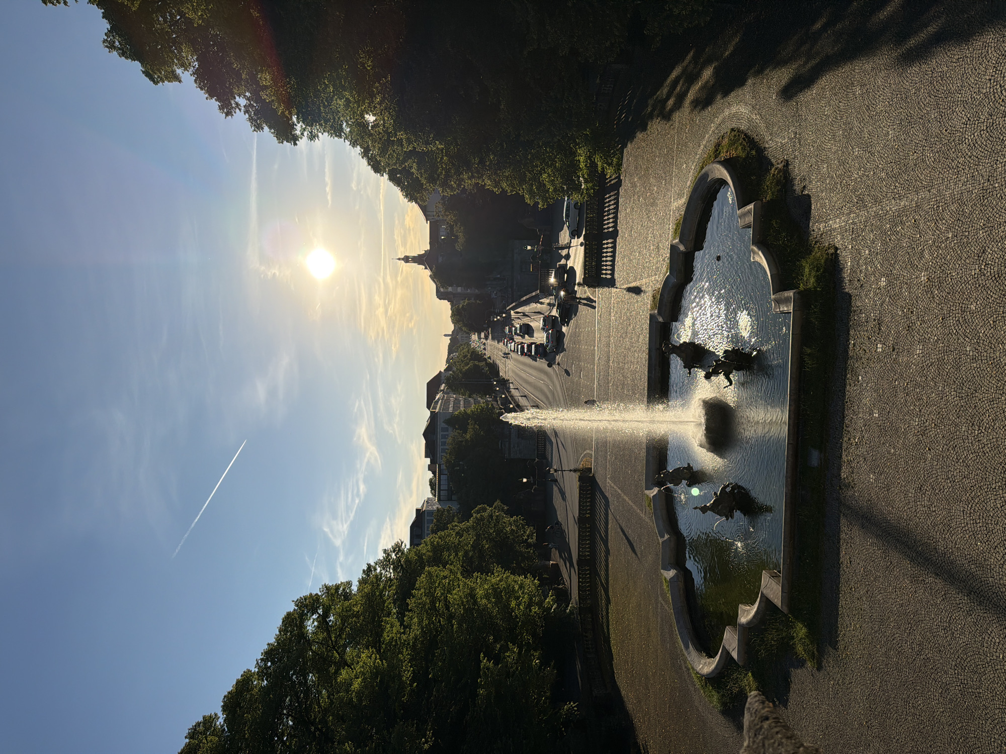 The image depicts a view from a high vantage point looking down a wide, tree-lined avenue in Munich, Germany, bathed in the warm glow of the setting sun. In the foreground, a circular fountain with sculpted figures is active, with water spraying upwards. The fountain is set within a paved area. Beyond the fountain, the avenue stretches into the distance, lined with cars and buildings. The sky is blue with wispy clouds and a clear contrail from an airplane. The sun is low on the horizon, casting long shadows. The environment appears to be a public space, likely a park or plaza, within a European city.