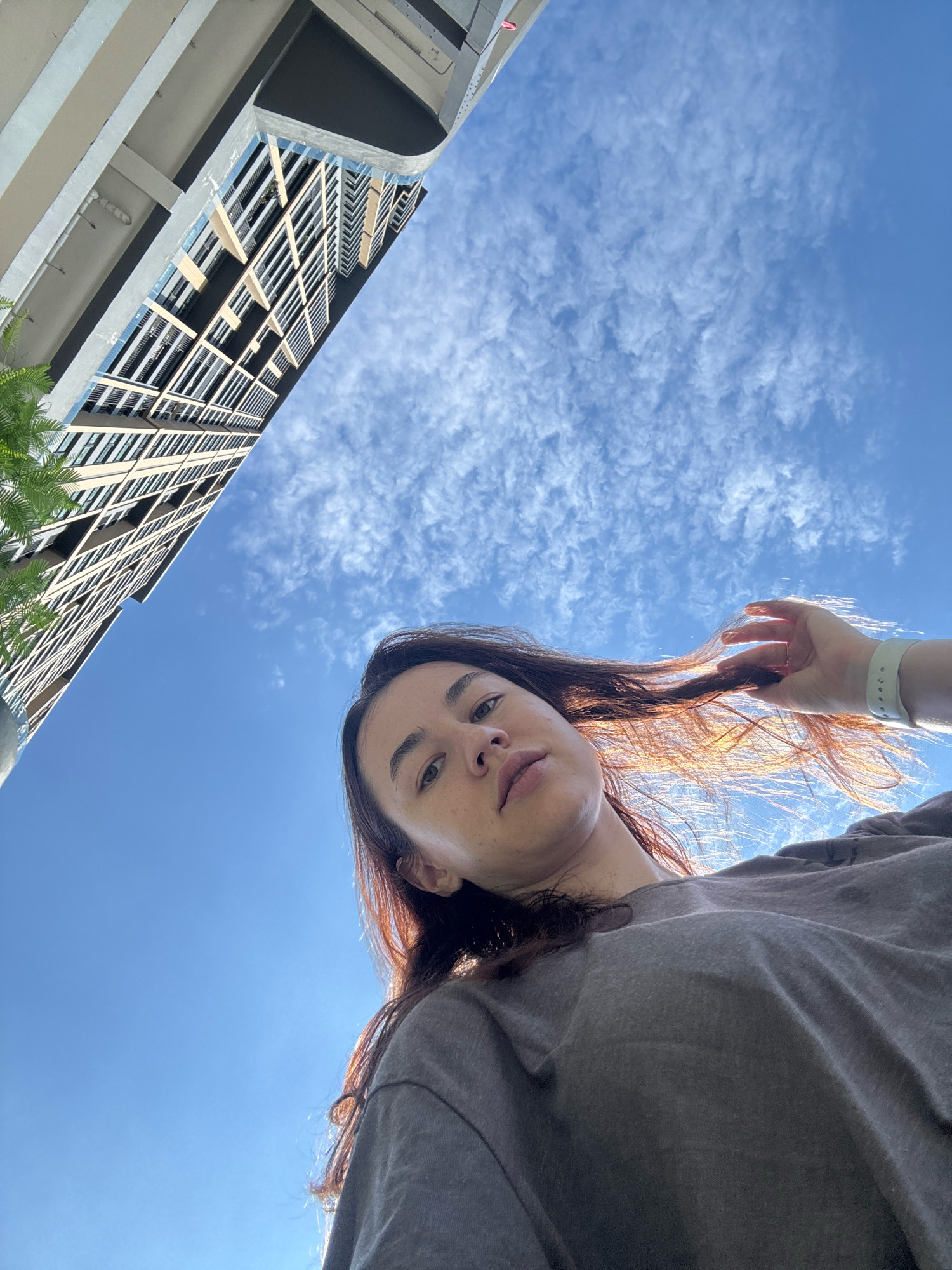 The image shows a low-angle, upward-facing shot of a young woman from the chest up, her face partially visible on the left side, looking towards the camera. Her long, dark hair is catching the sunlight, with some strands being touched by her right hand, which is adorned with a light-colored fitness tracker. She is wearing a grey t-shirt. In the background, a modern, multi-story building with numerous windows is visible, angled upwards. The sky is a clear blue with scattered white clouds. The scene appears to be outdoors in an urban environment, possibly within the vicinity of Samrong Nuea Subdistrict Municipality, Thailand, given the architectural style of the building.