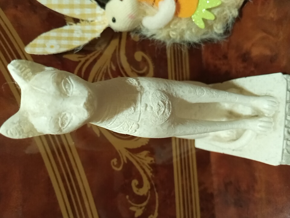 A white statue of a cat, likely an Egyptian Mau, is placed on a wooden table with an ornate, patterned surface. The cat statue is stylized, with distinctively large ears and almond-shaped eyes. It is in a seated position, with its front paws extended forward and its tail curled around its body. The statue has detailed markings on its body, including what appears to be a collar with a circular emblem in the center. Behind the cat statue, a portion of a decorative object with fabric ears, possibly part of a stuffed toy or decoration, is visible. The background is out of focus, but suggests an indoor setting. There is no visible text in the image. The lighting suggests it could be daytime, but the specific time of day or weather conditions are not discernible. No people are present in the image.