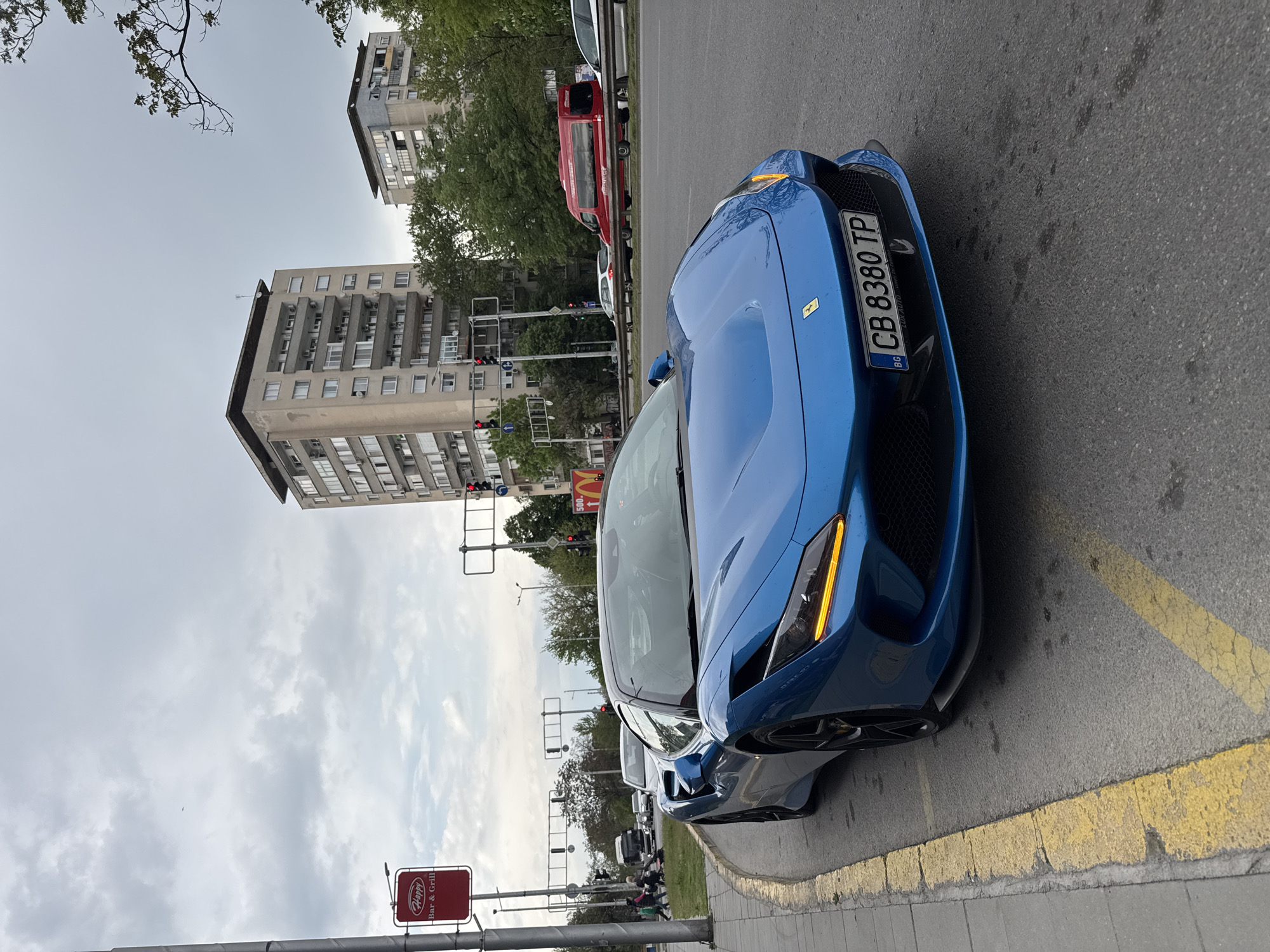 A vibrant blue sports car, appearing to be a Ferrari with its distinct badge on the front grille, is the prominent subject, positioned on the right side of an urban street. It is either parked or stopped by a yellow-painted curb marking. The car bears a Bulgarian license plate "CB 8380 TP" with "BG" below the characters. Its headlights have a distinct amber daytime running light.
The setting is an urban environment, identified as Plovdiv, Bulgaria. In the background, tall, multi-story buildings with numerous balconies and external air conditioning units line the street. The sky is overcast with a blanket of grey clouds.
Other vehicles, including a red van and a white car, are visible further down the road, suggesting ongoing traffic. Traffic lights are present at an intersection in the mid-ground. Small figures of pedestrians can be seen on the distant sidewalk.
Several signs are visible:
* A red rectangular sign with white text for "Happy Bar & Grill".
* A red sign with a yellow "M" logo for "McDonald's", with "500" underneath it.
The overall scene depicts a typical busy daytime street in a Bulgarian city under cloudy weather conditions.