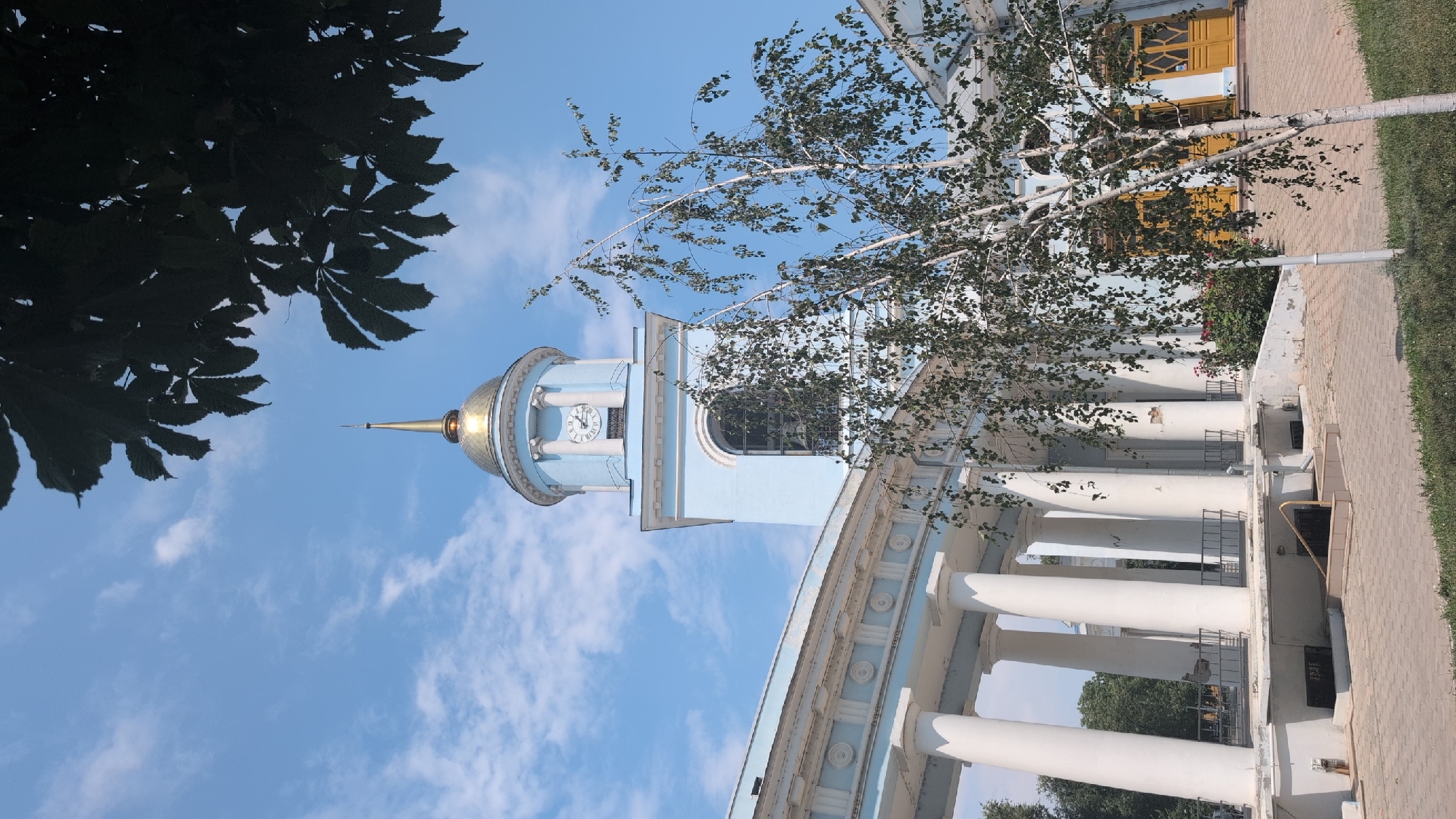 The image captures a view of the Izmail Holy Intercession Cathedral in Izmail, Ukraine. The cathedral features a light blue facade with white classical columns supporting an entablature. A prominent light blue tower with a golden dome and spire rises from the building, displaying a clock face. In the foreground, a slender birch tree with delicate leaves partially obscures the view of the cathedral's entrance. The ground is paved with light-colored rectangular tiles, and a strip of green grass runs along the bottom edge of the image. The sky above is a clear blue with scattered white clouds. There are no people visible in the scene. The overall impression is one of architectural grandeur against a bright, sunny day.