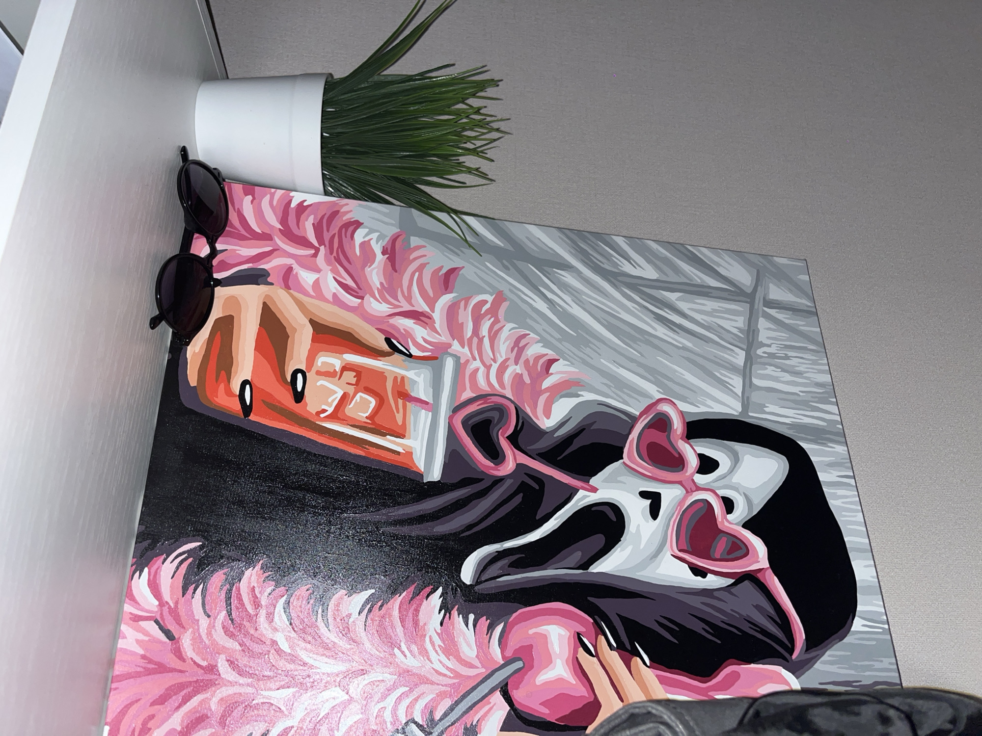 The image shows a painting propped against a wall or cabinet, with sunglasses hanging off the side. The painting depicts a cartoonish female figure with black hair and heart-shaped sunglasses. She's wearing a pink feather boa. There's a drink or object with rectangular shapes in the upper left corner of the painting. Above the painting is a white pot with green artificial grass. The wall behind is a light gray.
I am unable to determine if the location is Krasnodar, Russia from the information provided.