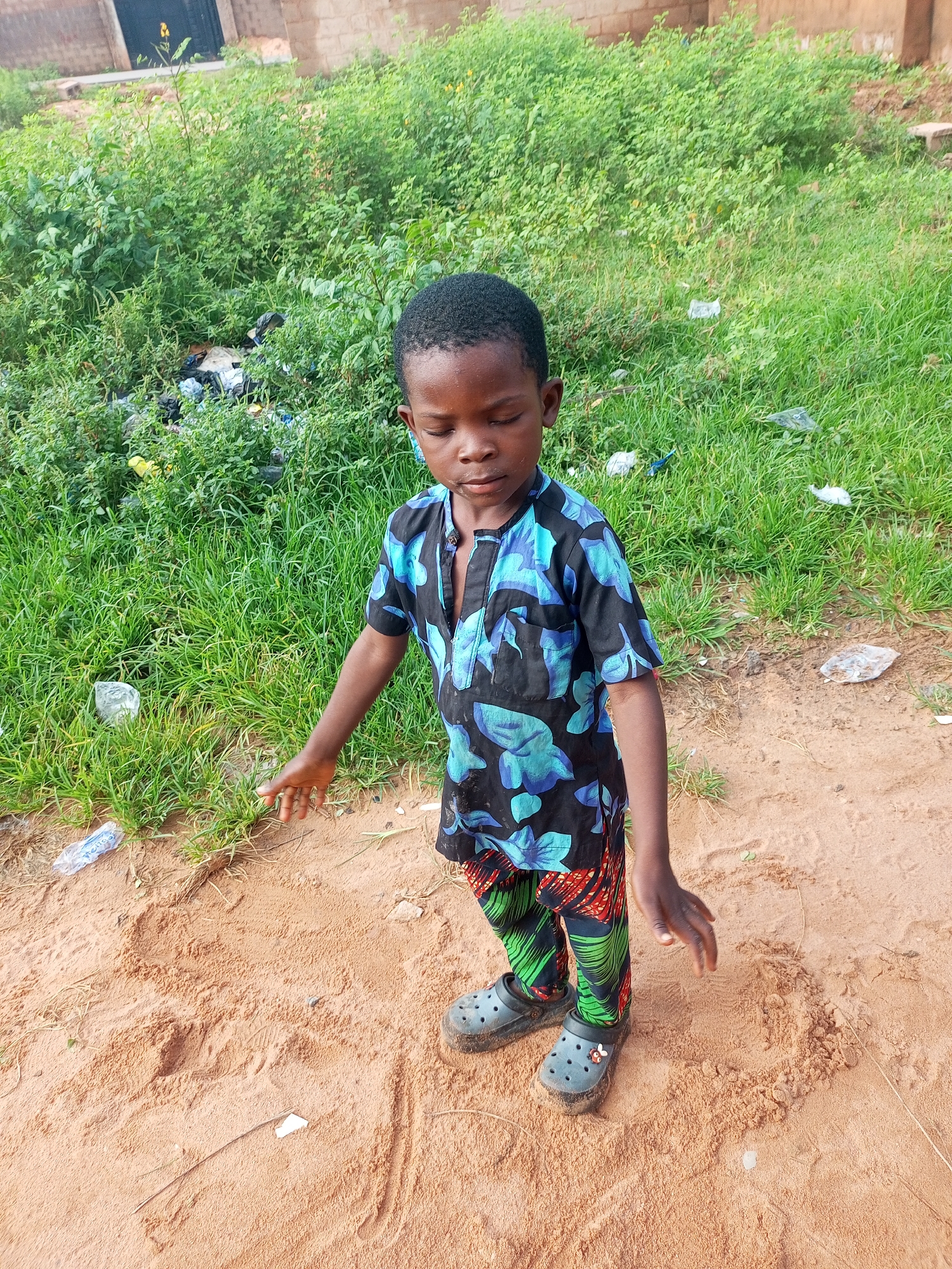 In a sandy area with sparse vegetation and some discarded waste, a young boy stands with his eyes closed and his head tilted down. He is wearing a black shirt with blue floral patterns and green and red patterned pants, along with dark grey crocs. His hands are slightly outstretched, and he appears to be in a moment of contemplation or play. The background consists of a dense growth of green plants and a building with a brick facade. This scene is set in Tempure, Nigeria.