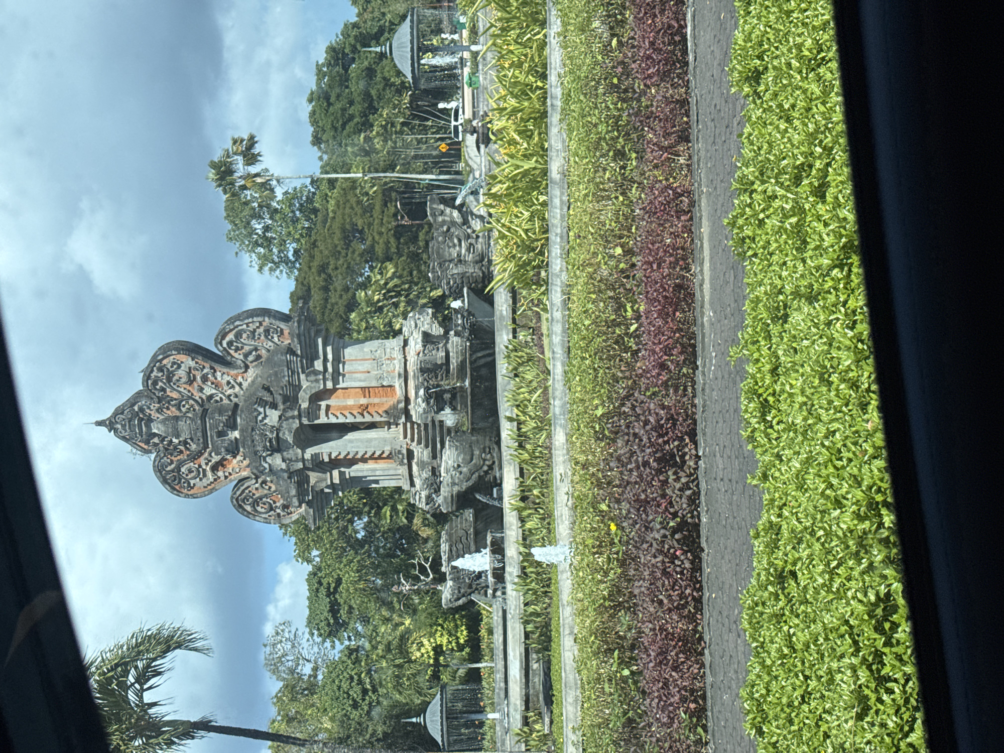 This image shows a view of the Dewa Ruci Roundabout in Kuta Selatan, Indonesia. A large, ornate stone monument, characteristic of Balinese architecture, stands in the center of the roundabout. It is surrounded by lush greenery and several fountains that are actively spraying water. The monument features intricate carvings and appears to be made of stone and brick. Palm trees and other tropical vegetation are visible in the background, along with a cloudy sky. The foreground is dominated by manicured bushes and a paved area, suggesting a public space or a road. No people are clearly visible in the image.