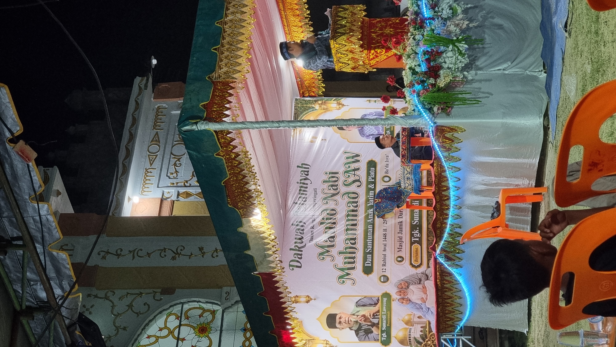 The image captures an outdoor evening event in Tumpokbeurandang, Indonesia, likely a religious gathering. A decorated stage with a banner reading "Dakwah Ramah Tamiyah" and "Maulid Nabi Muhammad SAW" is set up. On the stage, a man in a dark patterned shirt and cap is speaking into a microphone from behind a podium adorned with red flowers. To his left, another man is seated. A banner behind them features portraits of individuals and text in Indonesian. In the foreground, several orange plastic chairs are visible, with a young boy sitting in one, looking towards the stage. The background reveals a building with stained glass windows and decorative elements. The overall atmosphere suggests a community event taking place at night, illuminated by stage lighting and decorative string lights.