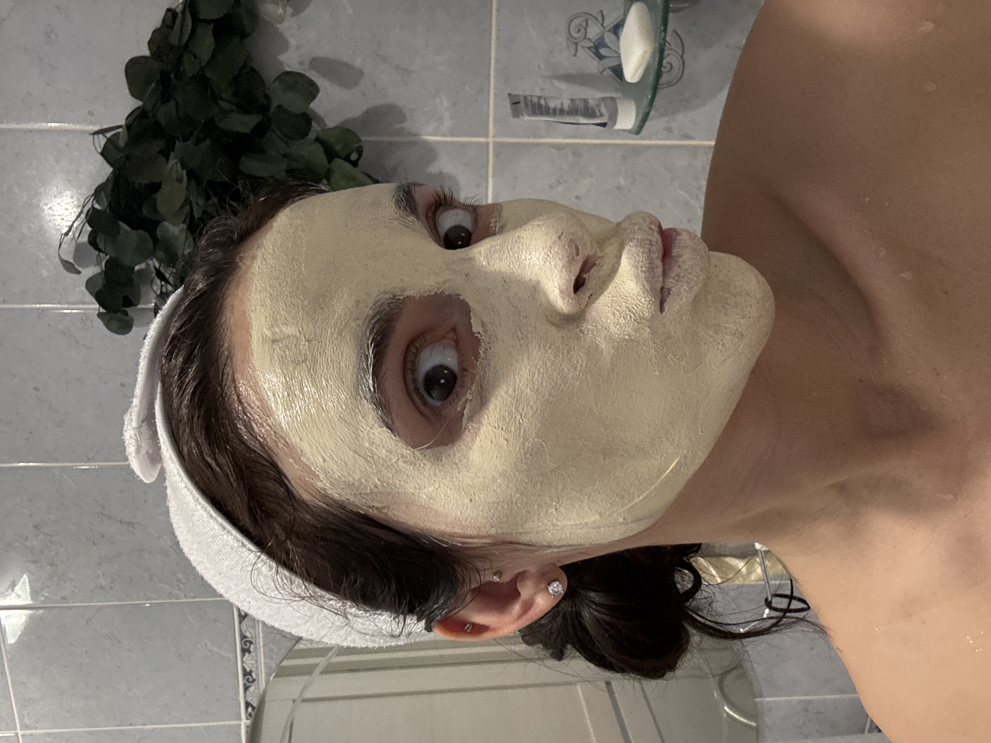 A woman with a face mask, white towel, and visible earrings is positioned in a bathroom setting. The bathroom features a tiled wall, plants, and bathroom accessories. The location is Chaikovsky, Russia.