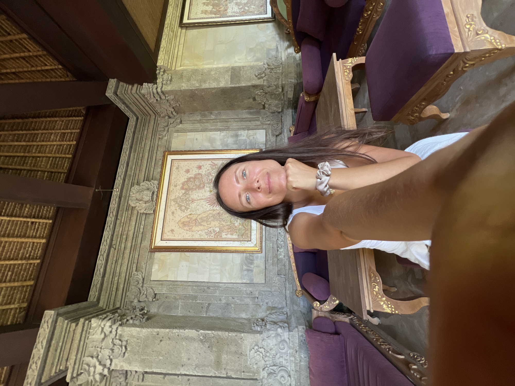 The image is a selfie taken indoors, likely within a building in Kedewatan, Indonesia. A woman with dark hair and blue eyes is the primary subject. She wears a white top and has a scrunchie on her wrist. Her arm is extended towards the camera, creating the selfie perspective.
The background consists of ornate stone walls, decorative carvings, and framed artwork on the walls. There are purple cushioned chairs and wooden tables with ornate detailing. A dark wooden structure, possibly part of the building's architecture, is visible in the upper left corner, along with a thatched ceiling. The lighting is bright and natural.