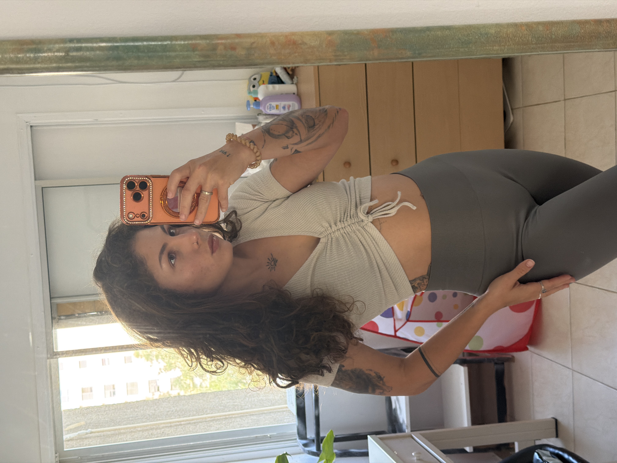 A young woman is captured in a mirror selfie, standing in a well-lit indoor space. She has long, wavy dark brown hair and an average build. She wears a light-colored, ribbed crop top with a central drawstring and dark grey high-waisted leggings. Visible tattoos include large designs on her arms, a small sun outline on her chest, and the text "I AM" on her left wrist, just below a coiled, bead-like bracelet. She also wears a ring on her right ring finger and has a nose piercing. The woman holds an orange smartphone with a decorative, possibly jeweled, case to take the photo, looking towards the camera with a calm, composed expression.
The setting appears to be a home, possibly a bedroom or living area. Behind the woman, to her right, a light brown wooden cabinet or chest of drawers is topped with baby care products, including what appears to be Johnson's Baby shampoo and lotion, and several small colorful toys. A colorful polka-dot laundry hamper or toy bin sits on the tiled floor to her right. To her left, a large window allows bright natural light into the room, revealing distant residential buildings under a clear sky, consistent with an urban or suburban landscape in Kiryat Bialik, Israel. The mirror she is using has a distinct, textured frame along its top edge. Some green plant leaves are visible in the bottom right foreground.