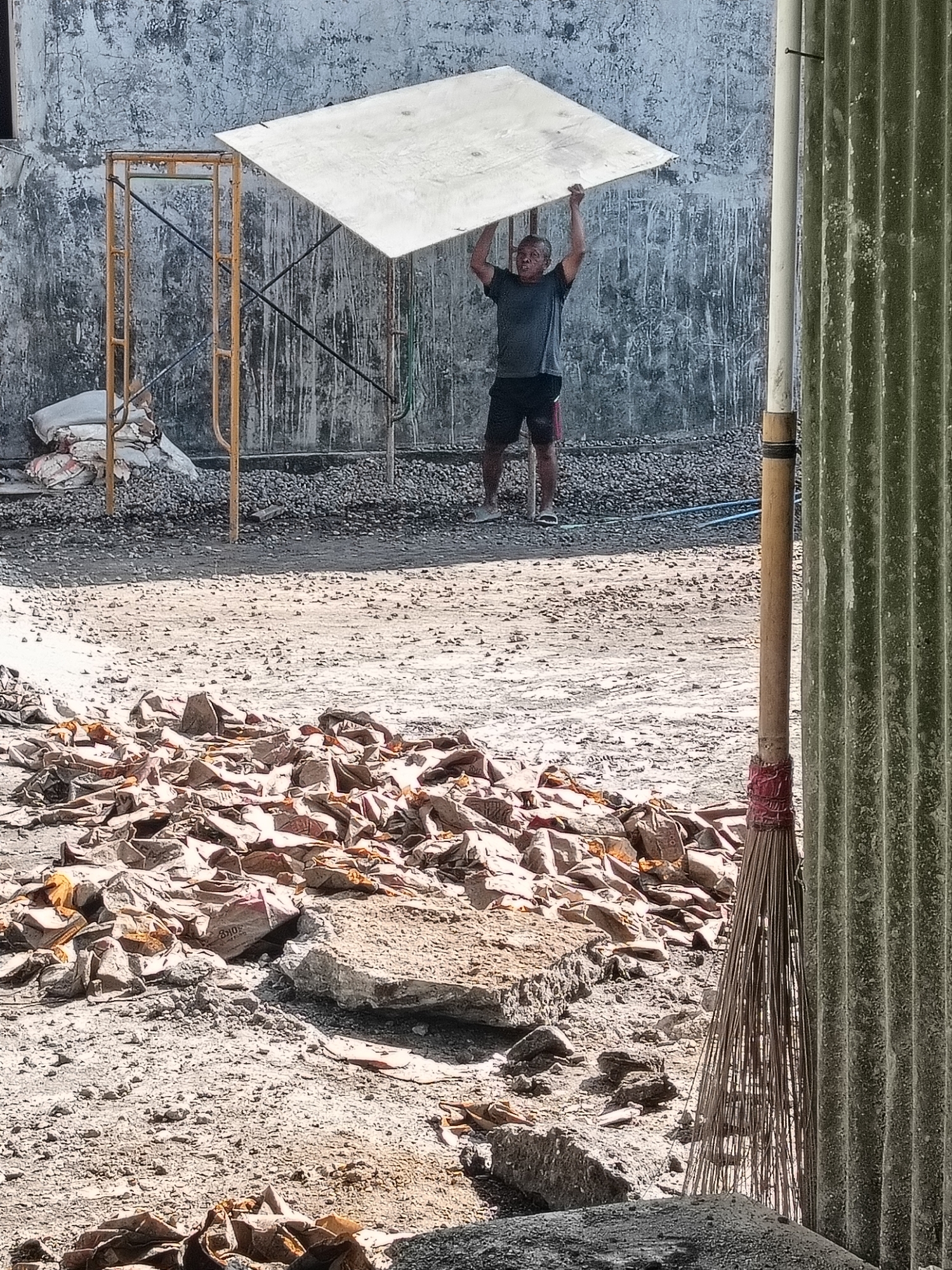 The image depicts a construction site in Gedangan, Indonesia. A man is holding a large, flat, light-colored object, possibly a piece of plywood or sheet metal, above his head. He is wearing a dark t-shirt and shorts, and appears to be on a construction project. The ground is covered in dirt, gravel, and debris, including broken bricks and what appear to be discarded bags. In the background, there is a concrete wall with a rough texture and some staining. Yellow scaffolding is visible to the left of the man, and a corrugated metal structure stands on the right side of the frame, partially obscuring a broom made of twigs. The overall scene suggests ongoing construction or demolition work.